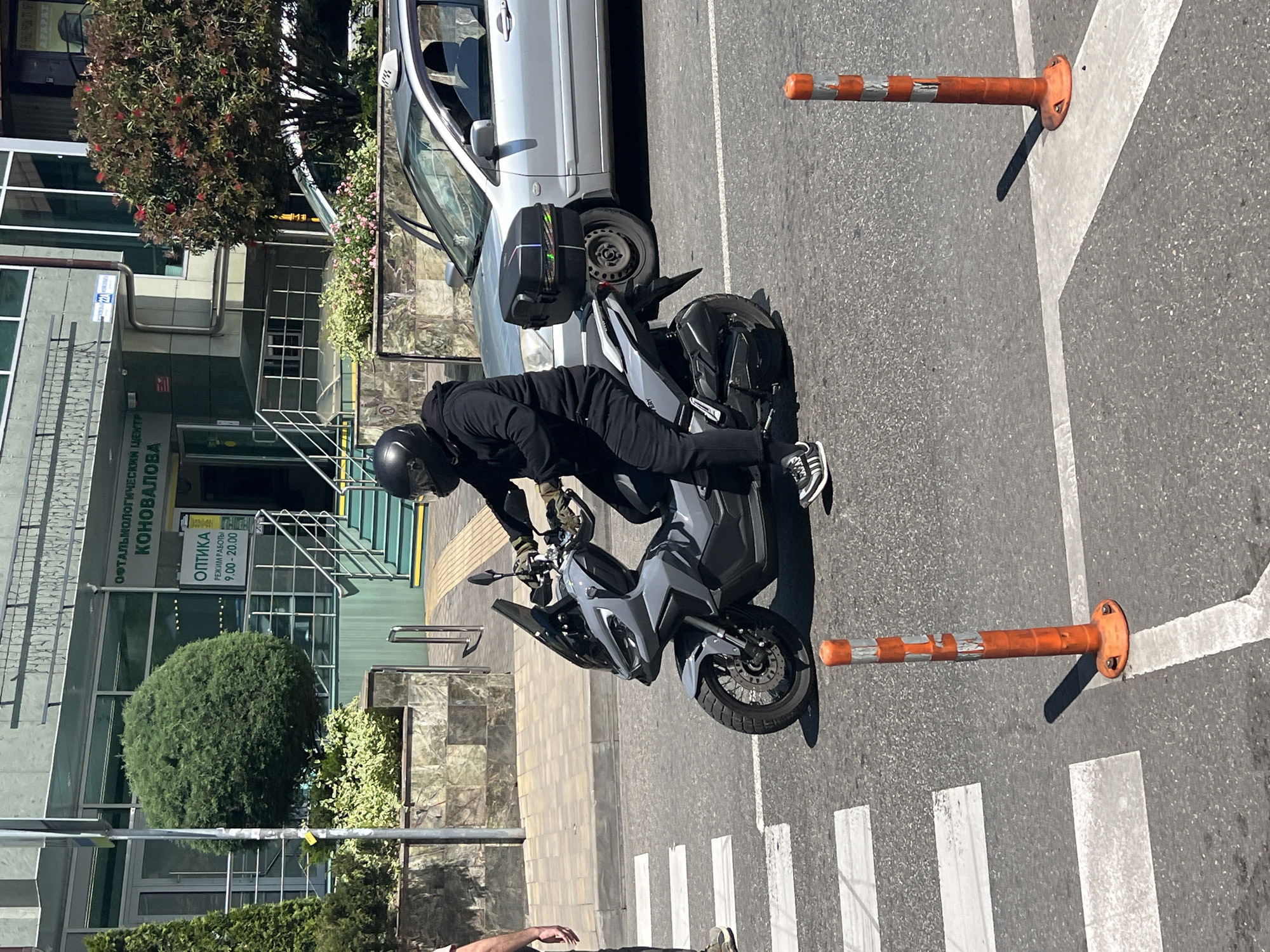 The image displays an urban street scene in Sochi, Russia, under bright daylight conditions, suggesting clear weather. The perspective is slightly elevated, looking down onto a street with an adjacent modern building.
In the center of the frame, a person wearing a black outfit and a black helmet is positioned on a grey and black scooter-style motorcycle. They appear to be stopped, leaning slightly forward, with feet near the ground. The motorcycle is equipped with a black storage case at the rear. On the asphalt road, two orange and silver flexible street bollards stand upright, marking the edge of a white pedestrian crosswalk or a designated area. White lines forming the crosswalk are clearly visible on the dark grey road surface. The arm and hand of another individual are partially seen in the lower left corner.
Behind the motorcycle, a silver sedan is parked alongside the curb. To the left, a multi-story building with a sleek glass and grey facade dominates the background. Its entrance features a staircase with metal railings. The area around the building is landscaped with green foliage, including manicured bushes and a tree adorned with red blossoms.
Visible text on the building's facade, in Cyrillic, reads "ОФТАЛЬМОЛОГИЧЕСКИЙ ЦЕНТР КОНОВАЛОВА" (Ophthalmological Center Konovalova). Below this, a smaller sign indicates "ОПТИКА" (Optics) and states "РЕЖИМ РАБОТЫ 9:00 - 20:00" (Working Hours 9:00 - 20:00). The overall scene depicts a typical daytime urban environment.