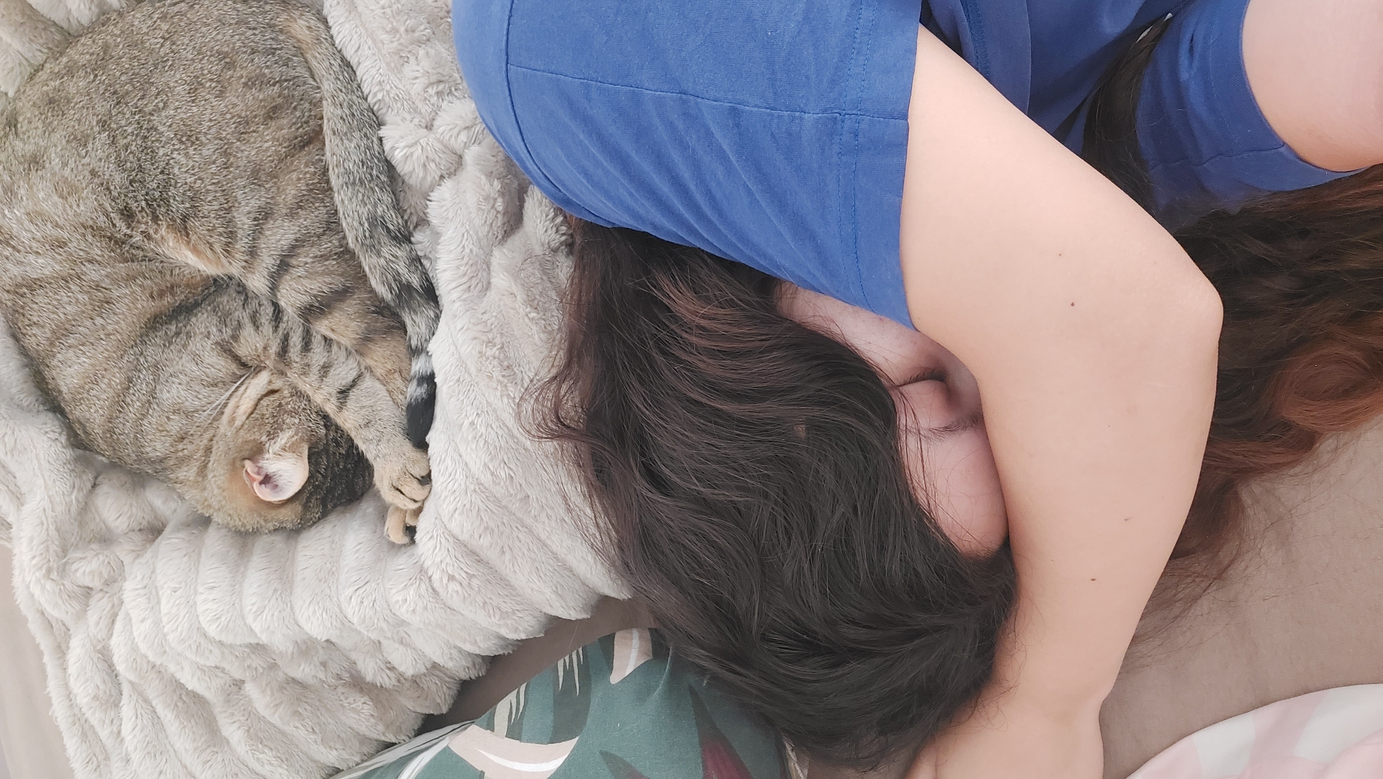 An indoor scene depicts a person and a tabby cat sleeping in close proximity. The person is positioned on the right side of the frame, lying on their side with their face partially obscured by their left arm. They have long, dark brown hair with visible reddish undertones, and are wearing a blue short-sleeved t-shirt. Their eyes are closed, indicating sleep. To the left and slightly above the person, a greyish-brown tabby cat with distinct darker stripes is curled tightly into a ball, also sleeping on a soft, light-colored, textured blanket. The cat's head is tucked in, with its left ear slightly visible, and its striped tail wraps around its body. The blanket covers what appears to be a bed or couch. A patterned pillow, featuring green, white, and dark red/brown abstract shapes, is visible on the far left, partially beneath the person's head. The lighting suggests a soft, ambient indoor setting. The geographical location, including the city of Shumen, Bulgaria, cannot be determined from the visual information presented in the image.