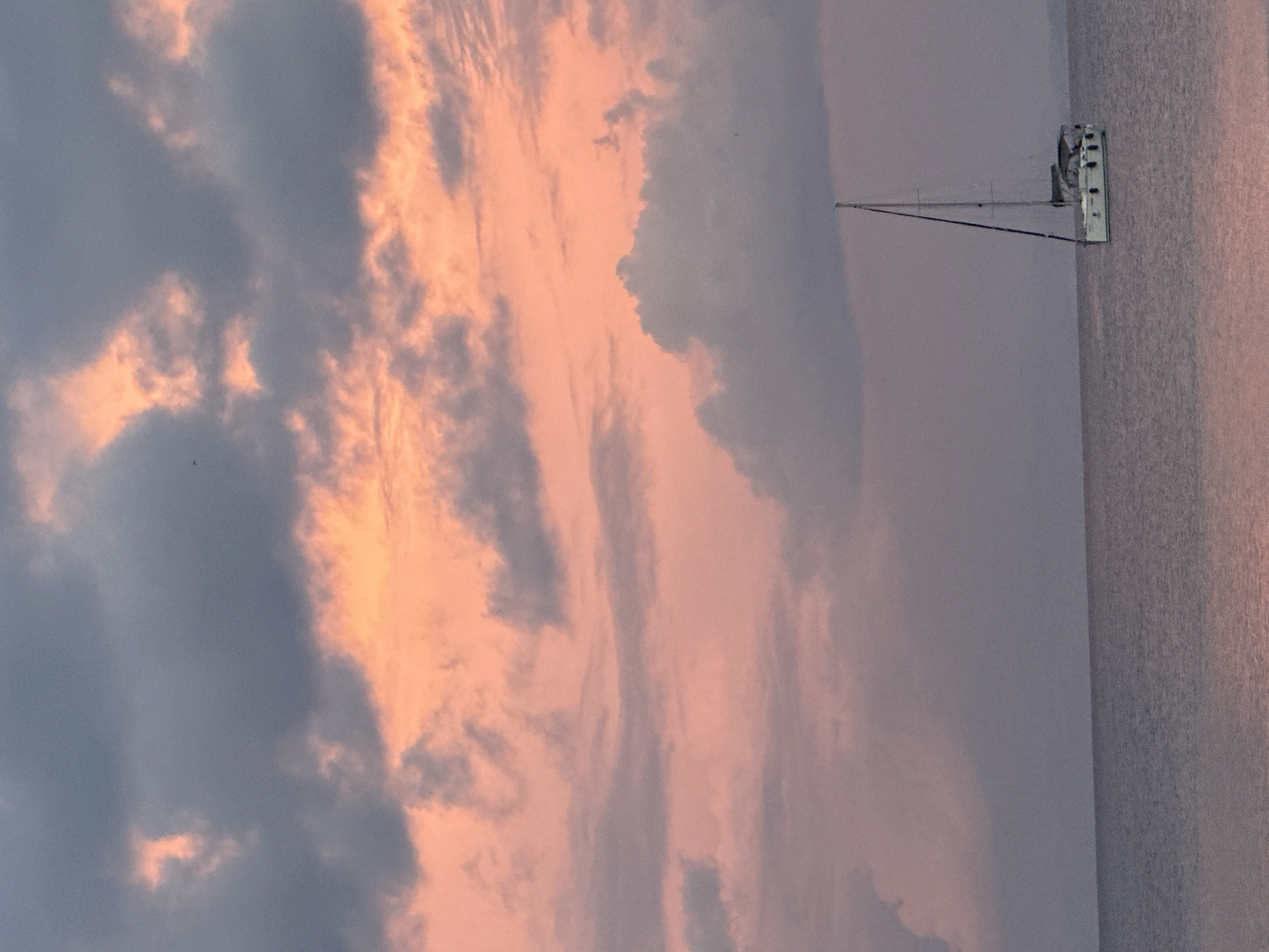 The image depicts a sailboat with its mast upright, floating on a calm body of water. The sky is filled with clouds, some tinged with shades of orange and peach, possibly indicating a sunset or sunrise. The overall tone of the image is serene and muted. Given the prompt's instruction, this scene may take place in Datça, Turkey.