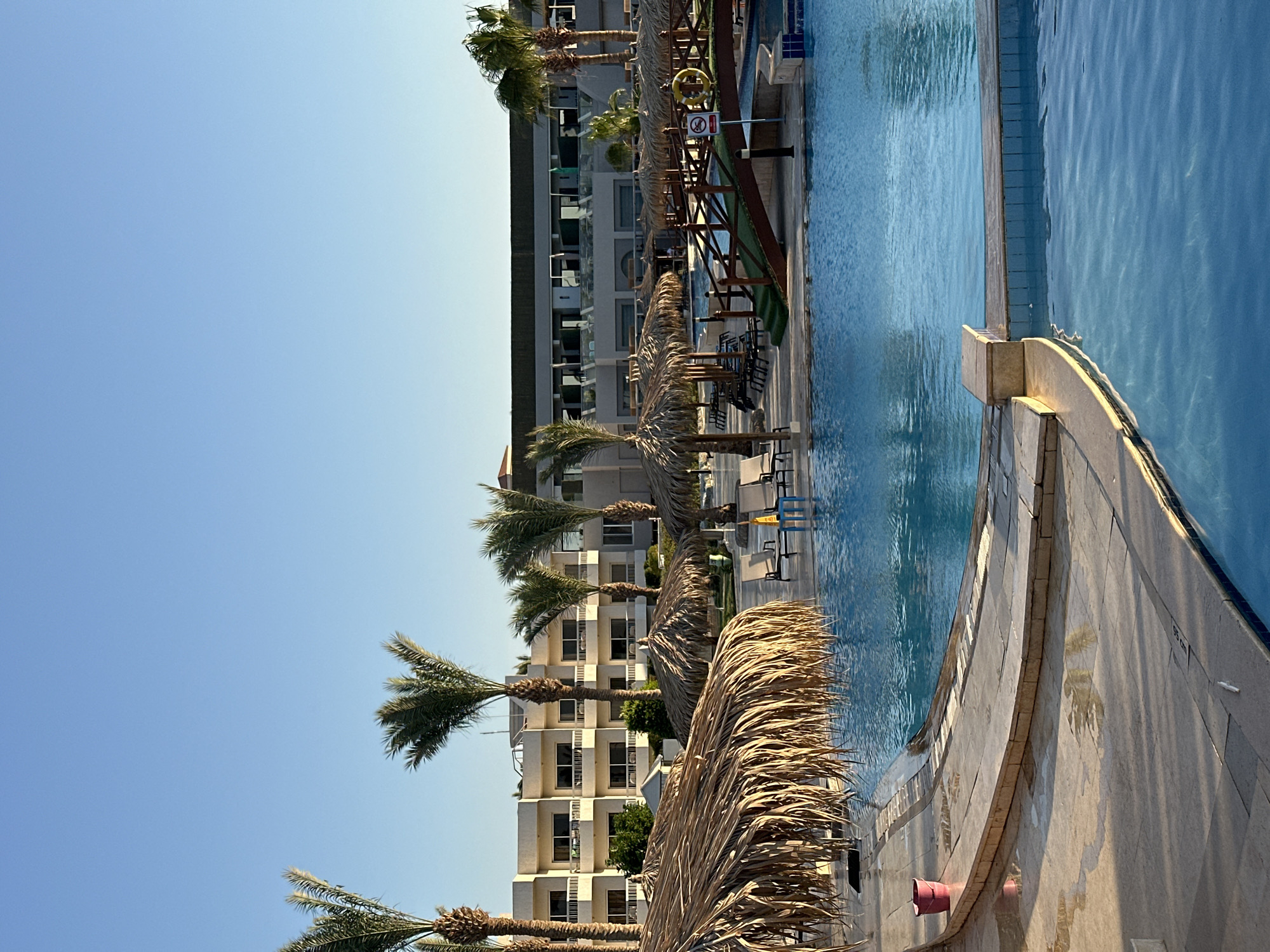 The image captures a poolside scene at a resort, likely in Hurghada, Egypt, given the presence of palm trees and the architectural style of the hotel buildings. The time of day appears to be late afternoon, as indicated by the warm, directional sunlight casting shadows. The weather is clear and sunny, with a bright blue sky dominating the upper portion of the frame.
The main subjects include a large, inviting swimming pool with clear blue water, a paved poolside area with lounge chairs and tables, and several palm trees with dry, straw-like fronds. In the background, a multi-story hotel building with balconies is visible, suggesting a resort or accommodation setting. There are no people actively engaged in the scene, creating a sense of quiet relaxation or anticipation.
Notable details include the thatched roofs of some poolside cabanas, providing shade to seating areas. The pool itself has a distinct edge with textured tiles and a decorative stone coping. Some light reflections are visible on the water's surface, and faint water marks or splashes are present on the poolside paving. The image is oriented vertically, with the sky at the top and the pool stretching downwards. The dry fronds of the palm trees suggest a warm, possibly arid climate, consistent with Hurghada. There is a yellow life preserver and a red sign with a crossed-out symbol (likely indicating a restriction) visible on a wooden structure near the pool. A measurement marking of "95 cm" is visible on the poolside paving. Two red buckets are also present near the edge of the pool area.