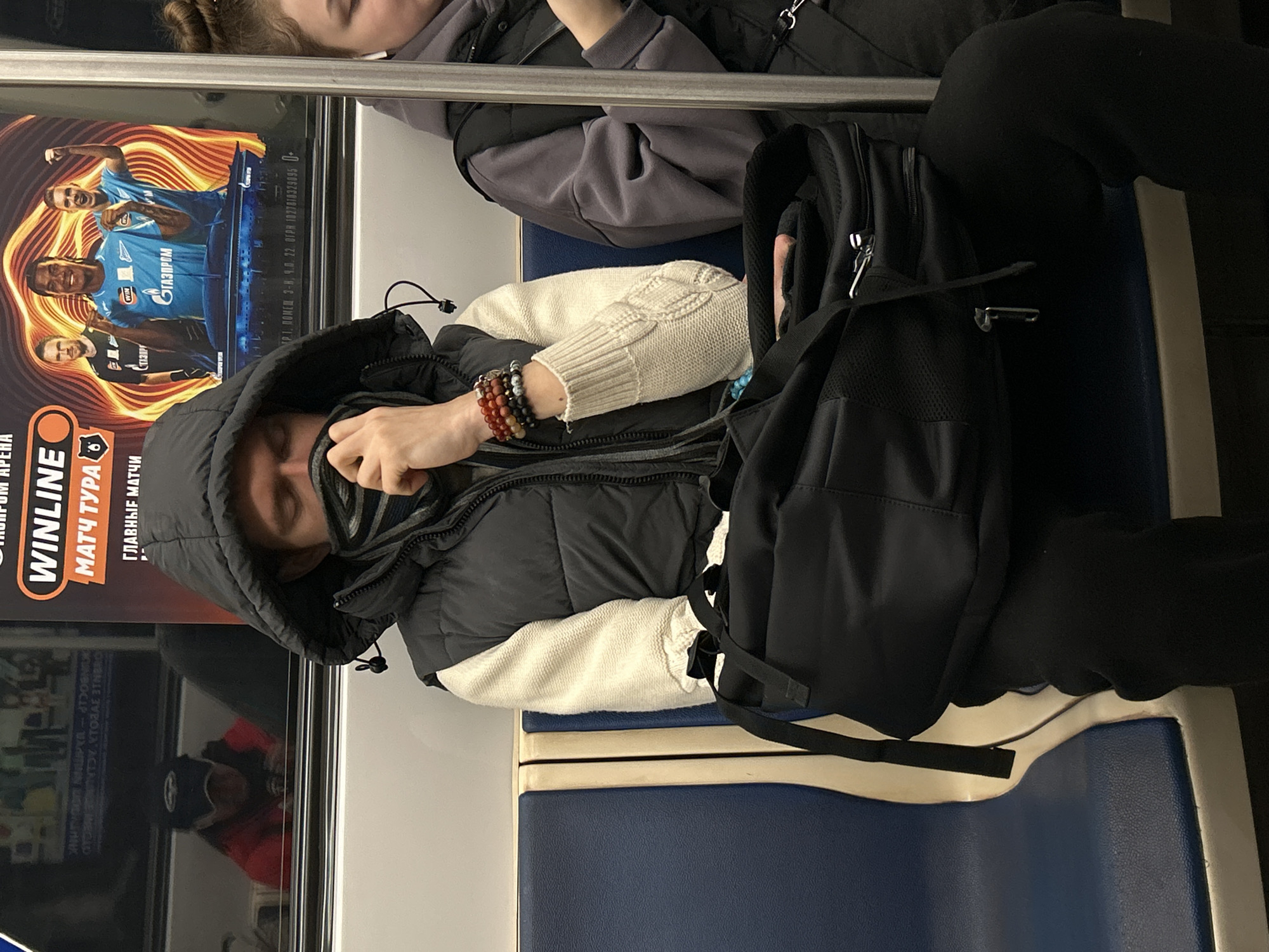 The image shows a person sitting on a blue and white seat in what appears to be a public transportation vehicle, possibly a metro train, as there's an advertisement next to the seat. The person is wearing a hooded jacket, a light-colored cable-knit sweater, and a patterned scarf covering part of their face. Their hand is positioned near their face, and several bracelets are visible on their wrist. A black backpack is placed next to them on the seat. Another person is partially visible in the upper portion of the image, seated next to them. The advertisement has the word "WINLINE" and features soccer players. Based on the advertisement content and the OCR, there's mention of "GAZPROM" and "Saint Petersburg," suggesting the location is in Saint Petersburg, Russia.