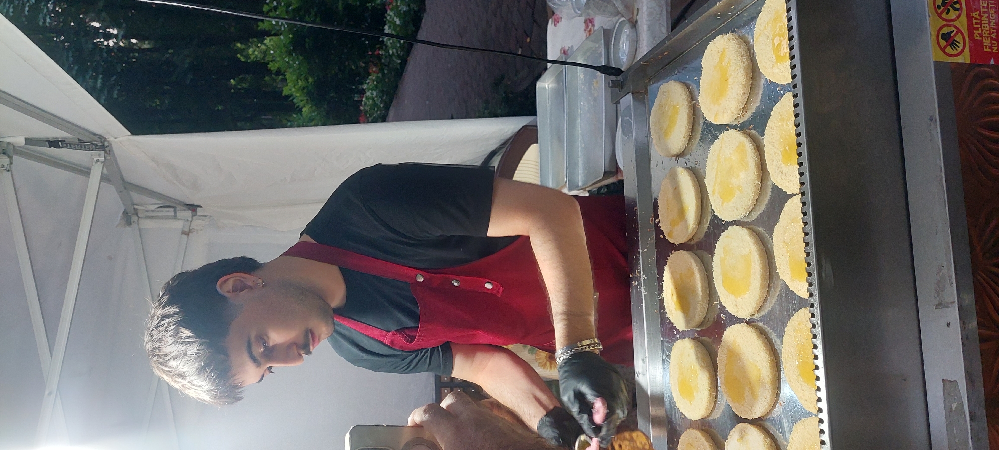 In Iași, Romania, a young man wearing a black t-shirt and a red apron is preparing food outdoors under a white tent. He has dark hair styled in a mullet and a mustache, and he is wearing black gloves. He is holding a phone in his left hand and appears to be transferring something from a container to a flat, round food item on a metal tray. The tray holds several similar food items, each coated in breadcrumbs and topped with a yellow substance, likely a sauce or filling. In the background, foliage and a walkway are visible, suggesting a park or outdoor event setting. A sign with Romanian text, "PLITĂ FIERBINTE NU ATINGE," is partially visible at the bottom right, indicating a hot plate and a warning not to touch.