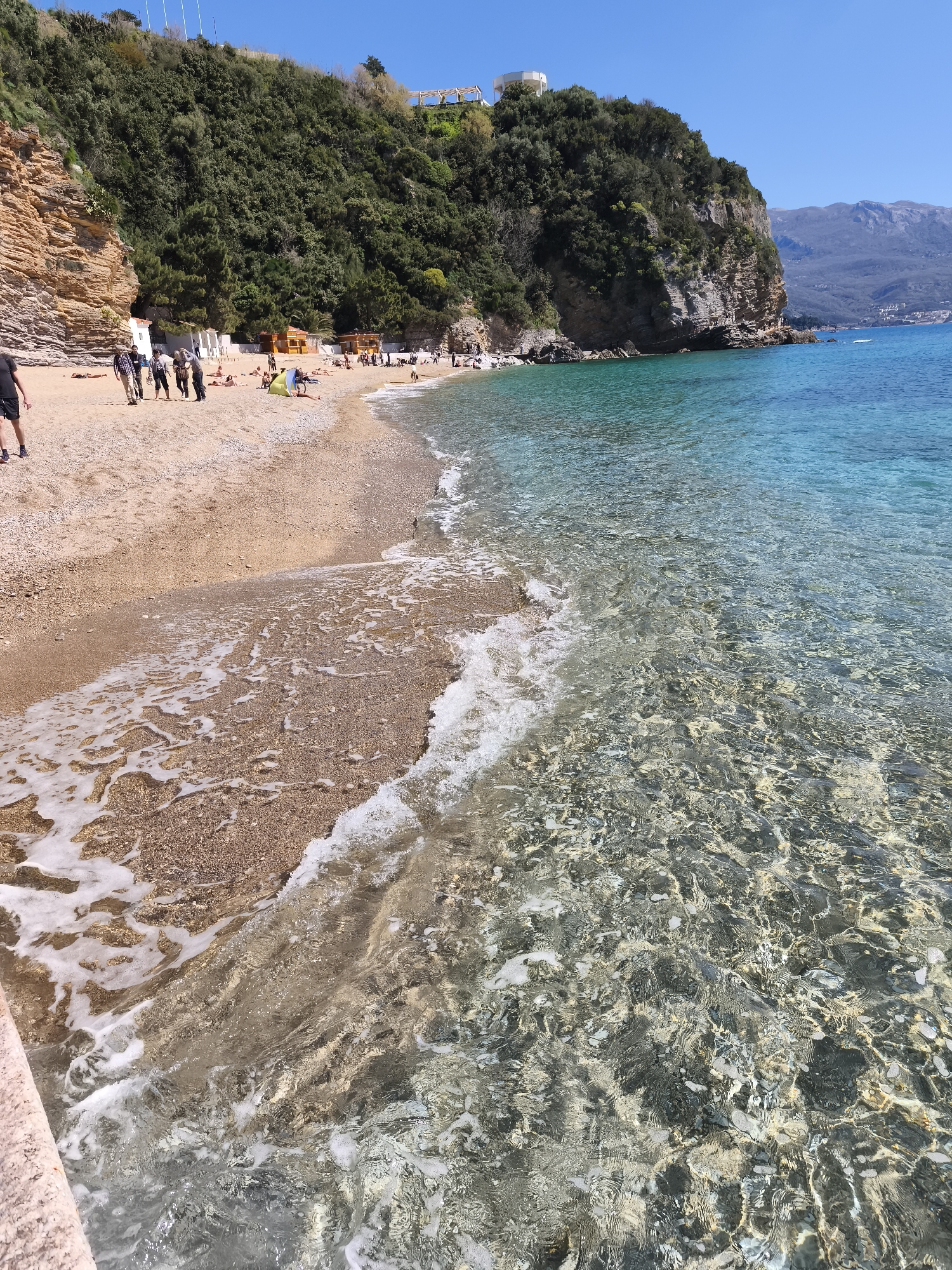 The image captures a sunny day at a beach in Budva, Montenegro. The foreground features crystal-clear turquoise water, with visible ripples and reflections. The shore is a mix of sand and small pebbles, with gentle waves lapping at the edge. Along the shoreline, there are several people, some walking, others relaxing on the sand. A small building or structure is on top of the rocky cliff behind the beach. Mountains are visible in the distance, adding to the scenic landscape. The vegetation is lush and green along the cliff.