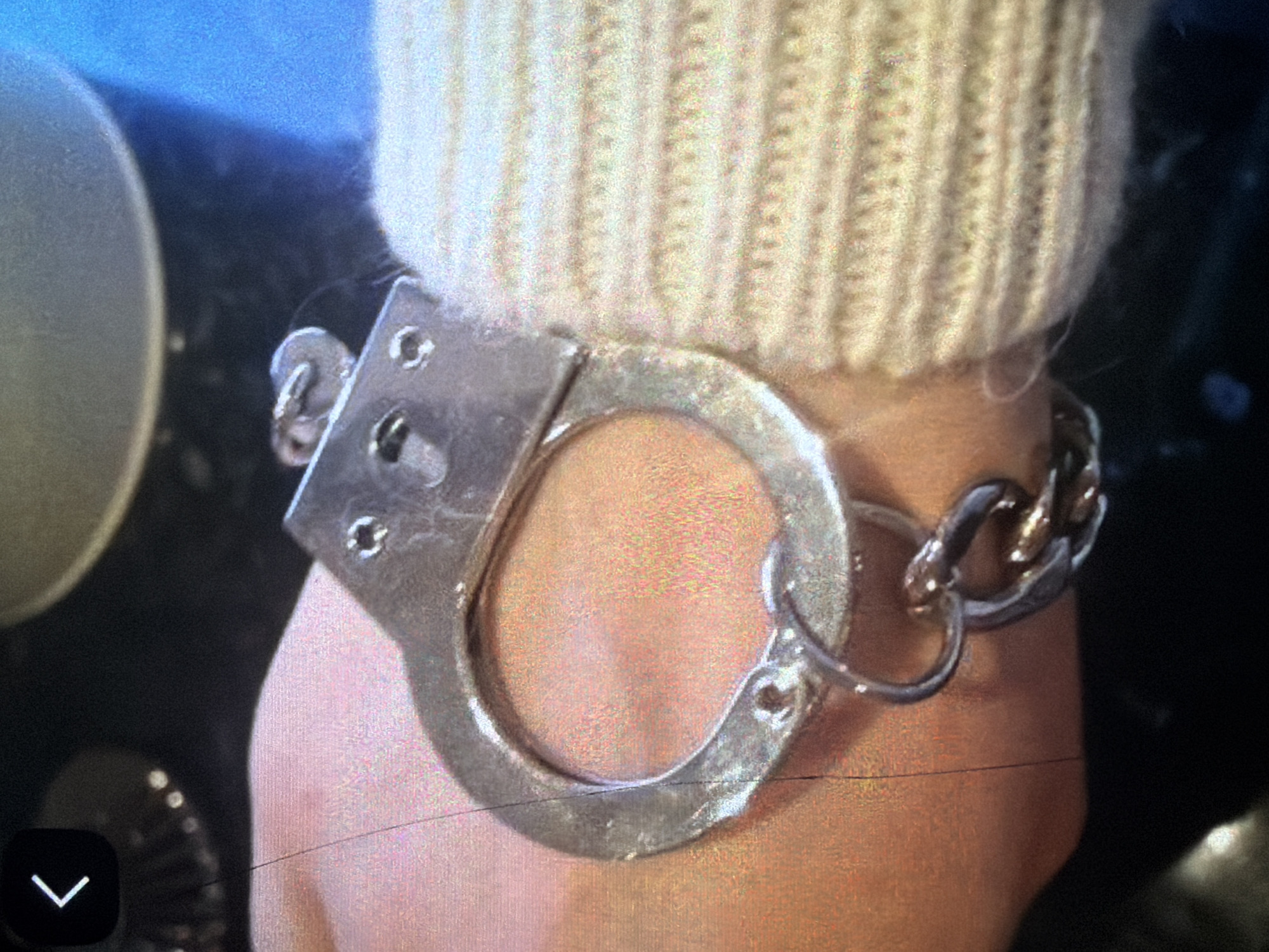 The image is a close-up shot of a person's wrist wearing a pair of metal handcuffs. The handcuffs are silver and appear to be of a standard design, with two interlocking cuffs connected by a chain. One cuff is fastened around the wrist, while the other is open. The person's skin is visible within the cuff, and they are wearing a beige knitted sweater on their arm. The background is dark and out of focus, with some blurred objects that suggest an indoor setting. There is no indication of the location being in Bukuru, Nigeria.