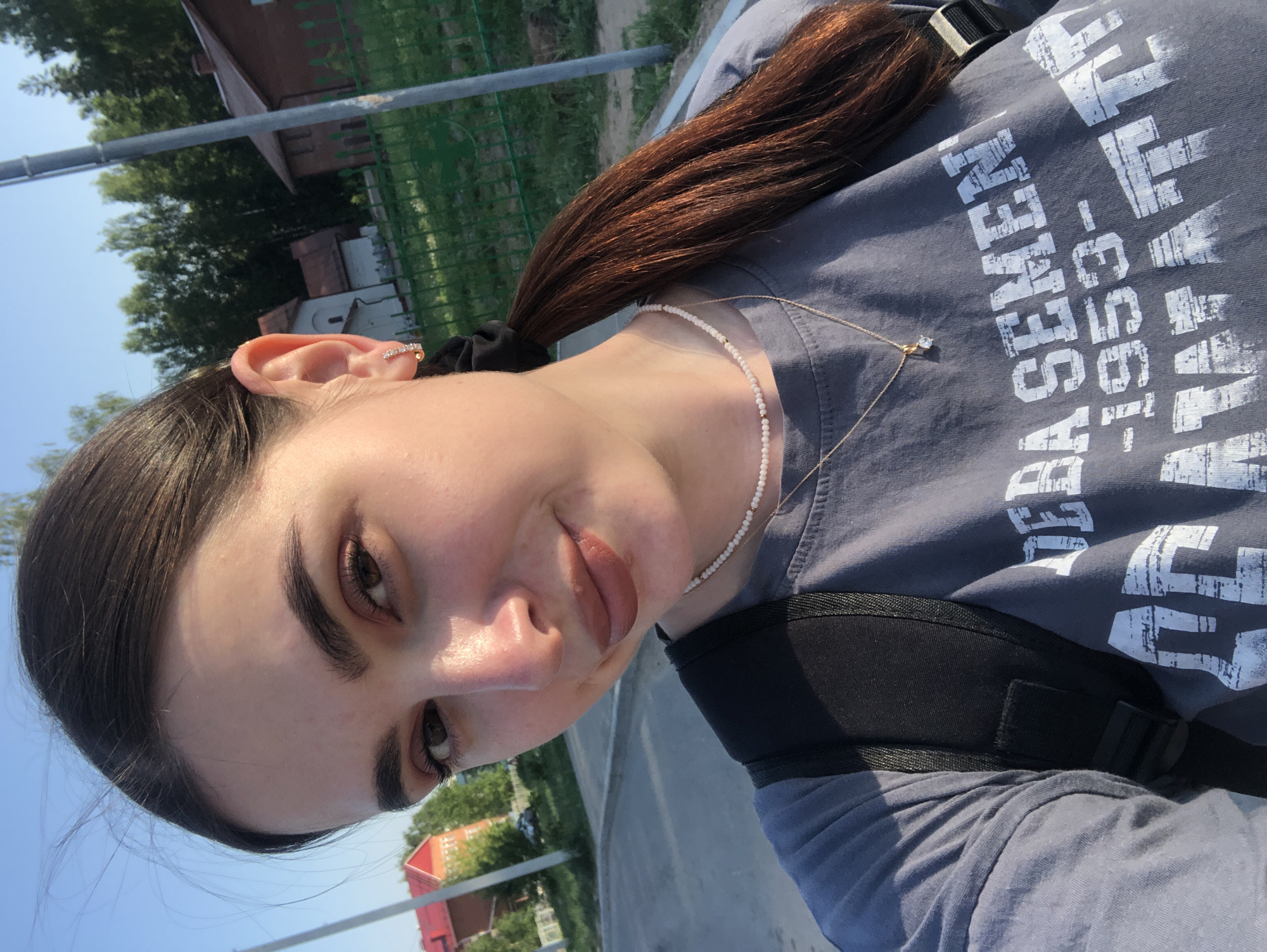 A young woman stands outdoors on a bright day. She is wearing a gray t-shirt with white lettering, a gold chain necklace, and a white beaded necklace. Her dark hair is pulled back in a ponytail secured with a black hair tie. She has a small earring in her upper ear. A black strap, possibly from a backpack, is visible across her chest. The background features buildings, trees, and a clear blue sky. Based on available knowledge, this image was taken in Noviy, Russia, where the specific location is indistinguishable.