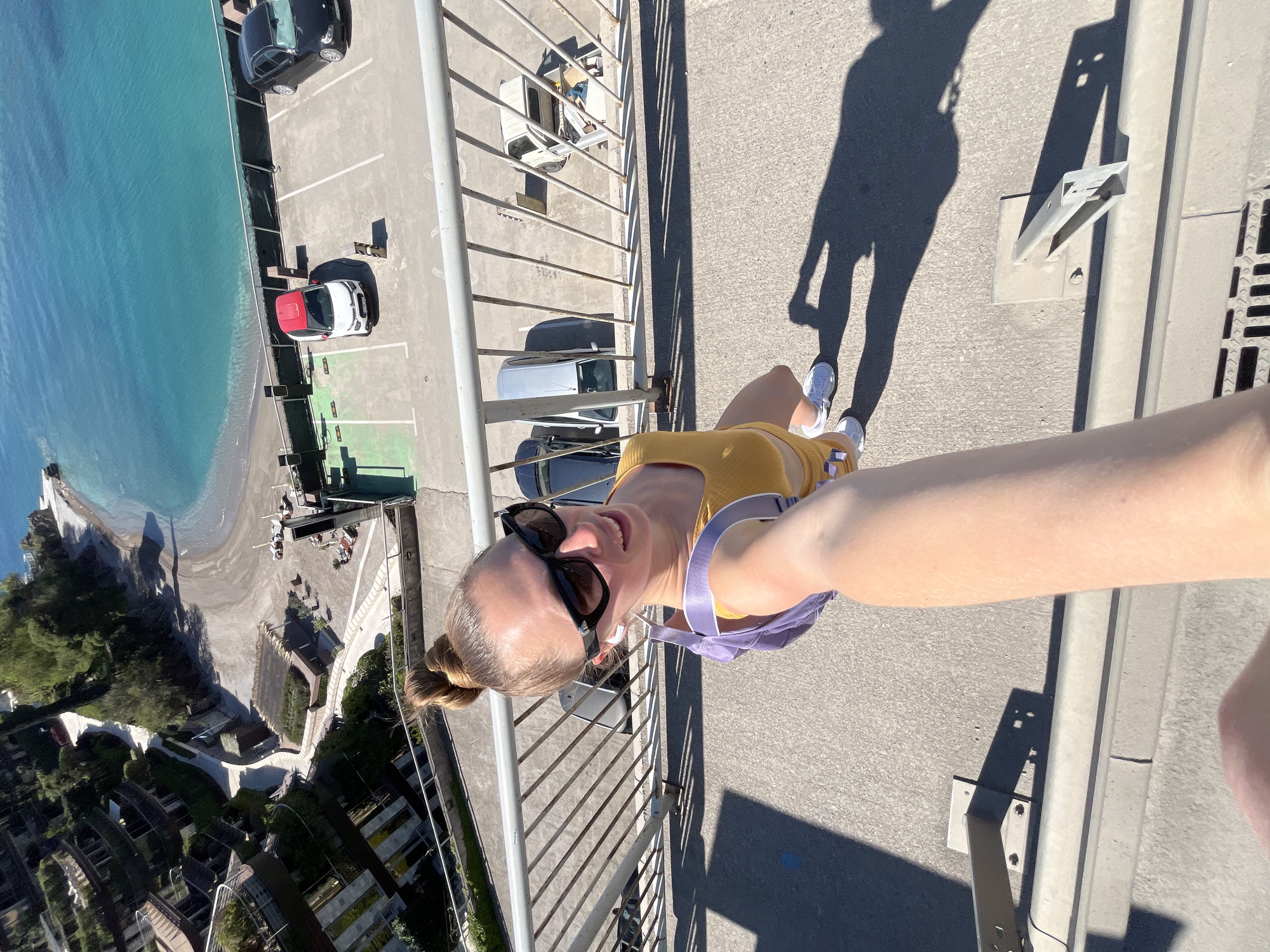 A rotated high-angle selfie features a woman standing on a bridge with a railing, overlooking a scene that includes a beach, buildings, and a parking lot. The woman is wearing sunglasses, a yellow outfit, and a purple bag. A partial shadow of the woman is visible on the bridge's surface, near the railing. In the background, a blue body of water borders a sandy beach. Adjacent to the beach is a parking lot with multiple vehicles parked. Beyond the parking lot, there are buildings and trees. The precise city is not discernible from the image.