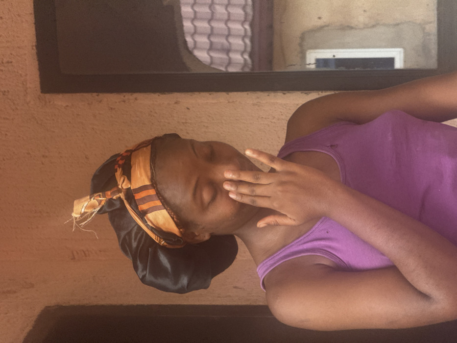 A woman reclines with her eyes closed, her right hand partially covering her face. She wears a purple tank top and a black satin bonnet with a patterned orange and brown headscarf tied around the edge. The background features a textured wall with a dark-framed window above. The angle of the shot is slightly tilted, giving a sense of informality.