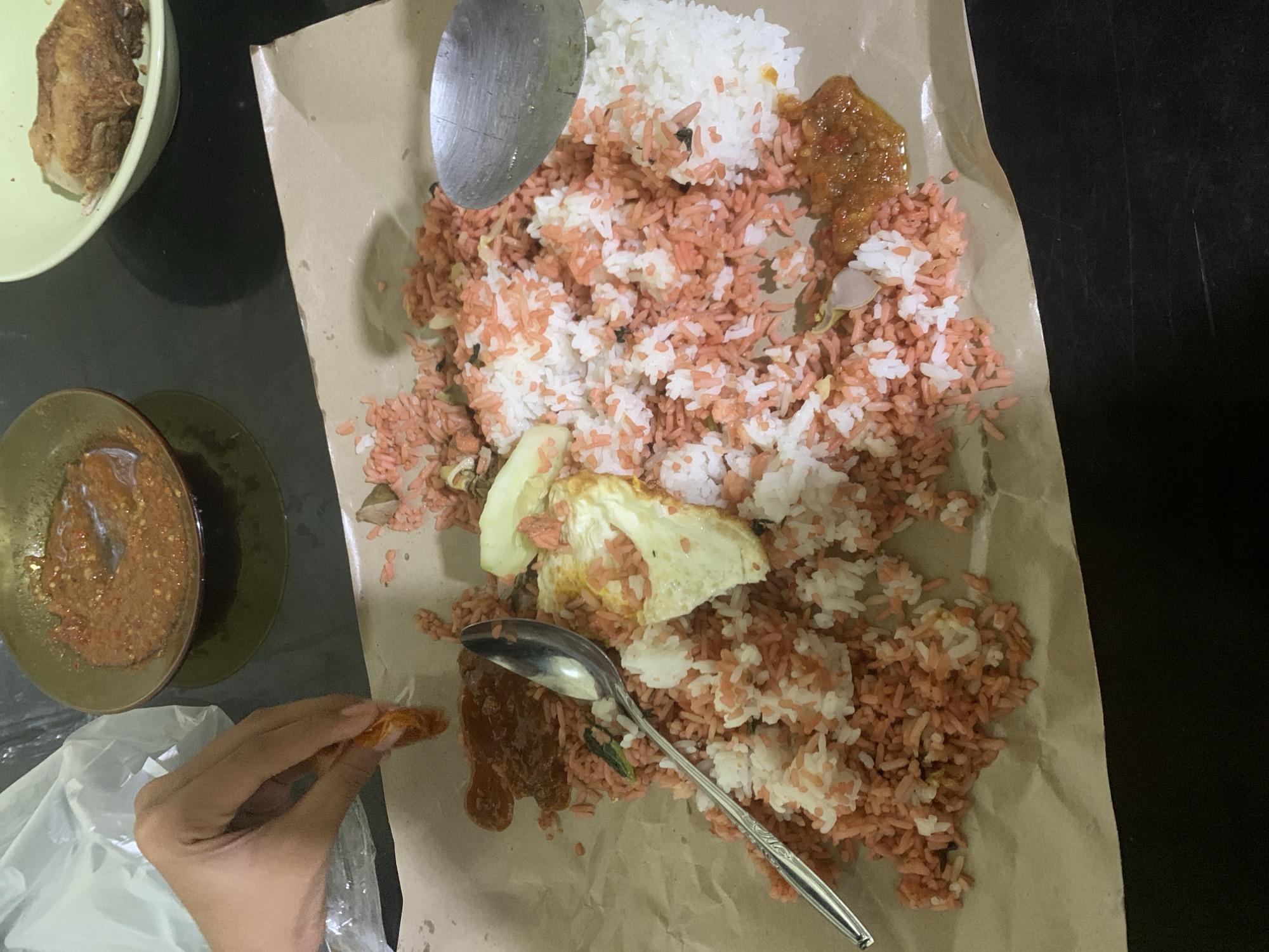 The image captures a close-up view of a meal served on brown paper, likely in Sungai Meriam, Indonesia. The meal consists of rice that is partially pink and partially white, mixed with small pieces of vegetables and what appears to be fried egg. A spoon is placed on top of the rice. To the left, a hand is visible, holding a piece of food and appearing to be in the process of eating. There are also two small bowls in the background, one containing a reddish sauce and another with a piece of fried food. The scene is set on a dark, reflective surface, possibly a table.