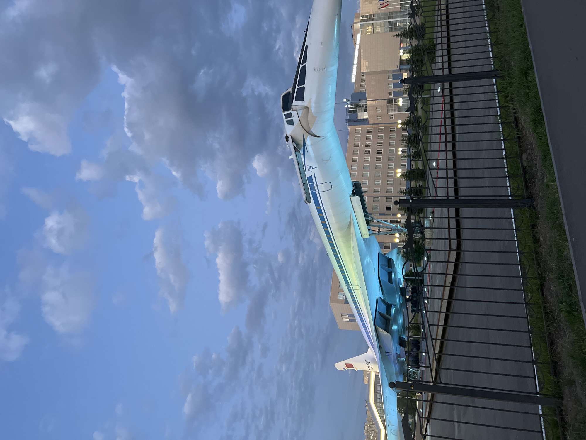 An outdoor photograph captures a Tupolev Tu-144 supersonic transport aircraft on display, illuminated by blue and green lights, in front of a modern building complex. The aircraft, painted white with blue and yellow stripes along its fuselage, has its nose section angled upwards. The registration CCCP-77107 is visible on the tail. The background features a multi-story building with numerous windows, and several evergreen trees are planted in front of it. A dark metal fence with decorative elements in the shape of airplanes runs across the foreground, separating the viewer from the aircraft. The sky above is filled with a mix of light and dark clouds. The scene appears to be set in the evening or dusk, with some lights visible in the building. The location is the Tupolev Tu-144 monument in Kazan, Russia. There are no people visible in the photograph.