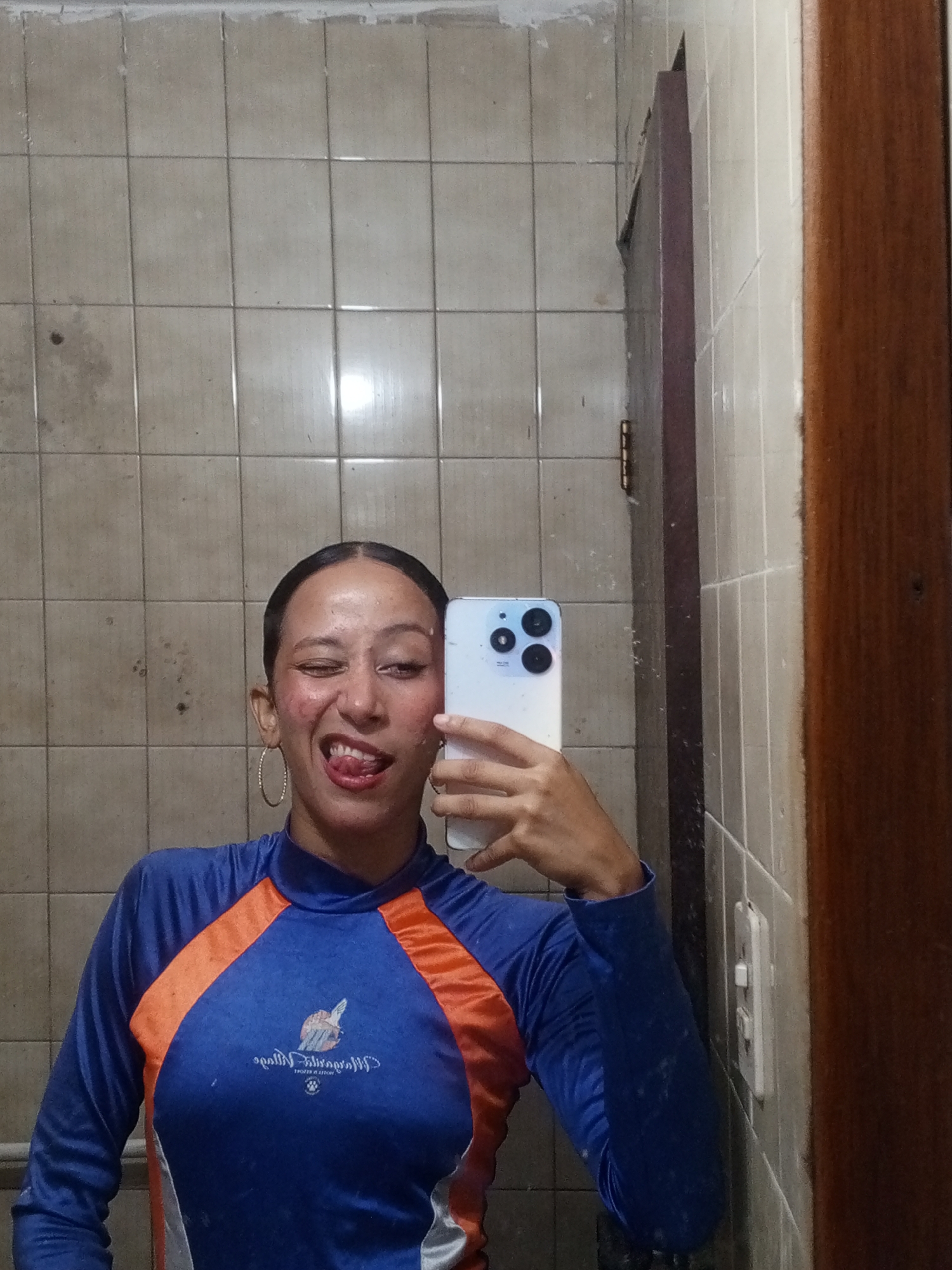 A young woman is depicted taking a mirror selfie in what appears to be a bathroom or changing area. She holds a light-colored smartphone with a distinctive camera module in her right hand, partially obscuring her face. Her dark hair is pulled back, and she wears a blue, long-sleeved rash guard or swim shirt. The shirt features bright orange panels on the shoulders and arms, and a white/grey stripe on the lower left side. She wears a large golden hoop earring on her visible left ear. Her expression is playful, with a wink of her left eye, her tongue slightly out, and a wide smile.
The background consists of light beige or off-white square tiles with dark grout lines. The tiles show signs of age and wear, with visible stains, smudges, and some dirt, suggesting a well-used environment. To her right, a dark brown wooden door frame is visible, along with a white light switch mounted on the tiled wall. A faint reflection of the door frame and switch can also be seen on the left side of the image.
On the front of her blue shirt, a logo and reversed text are visible. The logo appears to be a stylized creature, possibly a bird or phoenix, over wave-like shapes. Below the logo, the text reads "Miracle Village Hotel & Resort," with a small paw print graphic underneath. The image captures a candid moment in an indoor setting, likely in Porlamar, Venezuela.