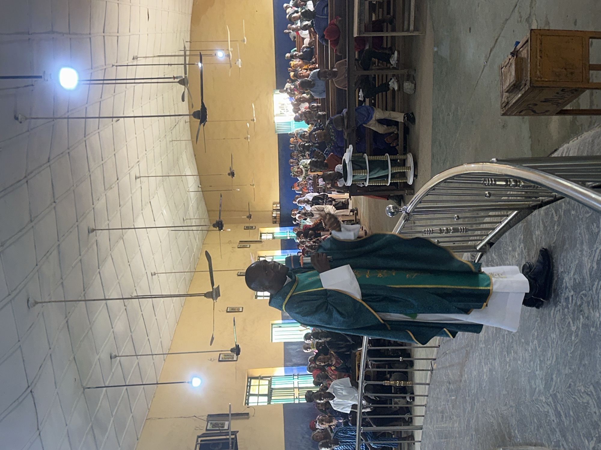 The image depicts a Catholic priest in vestments addressing a congregation gathered inside a church in Akwanga, Nigeria. The priest, a Black man wearing a green and gold chasuble over a white alb, stands on a raised platform to the left, holding a microphone in his right hand and gesturing with his left. The congregation, composed of men, women, and children of various ages, is seated on wooden benches in rows extending towards the back of the church. They are dressed in casual and traditional attire.
The church interior features a high, white, tiled ceiling with numerous ceiling fans and light fixtures. The walls are painted in alternating bands of yellow and blue. Large windows with green shutters are visible on the left side. The floor is tiled with a marbled pattern. In the foreground, a silver metal railing curves around the platform. To the right of the frame, a wooden collection box with a lid is placed on the floor. The overall atmosphere appears to be one of a religious service or gathering.