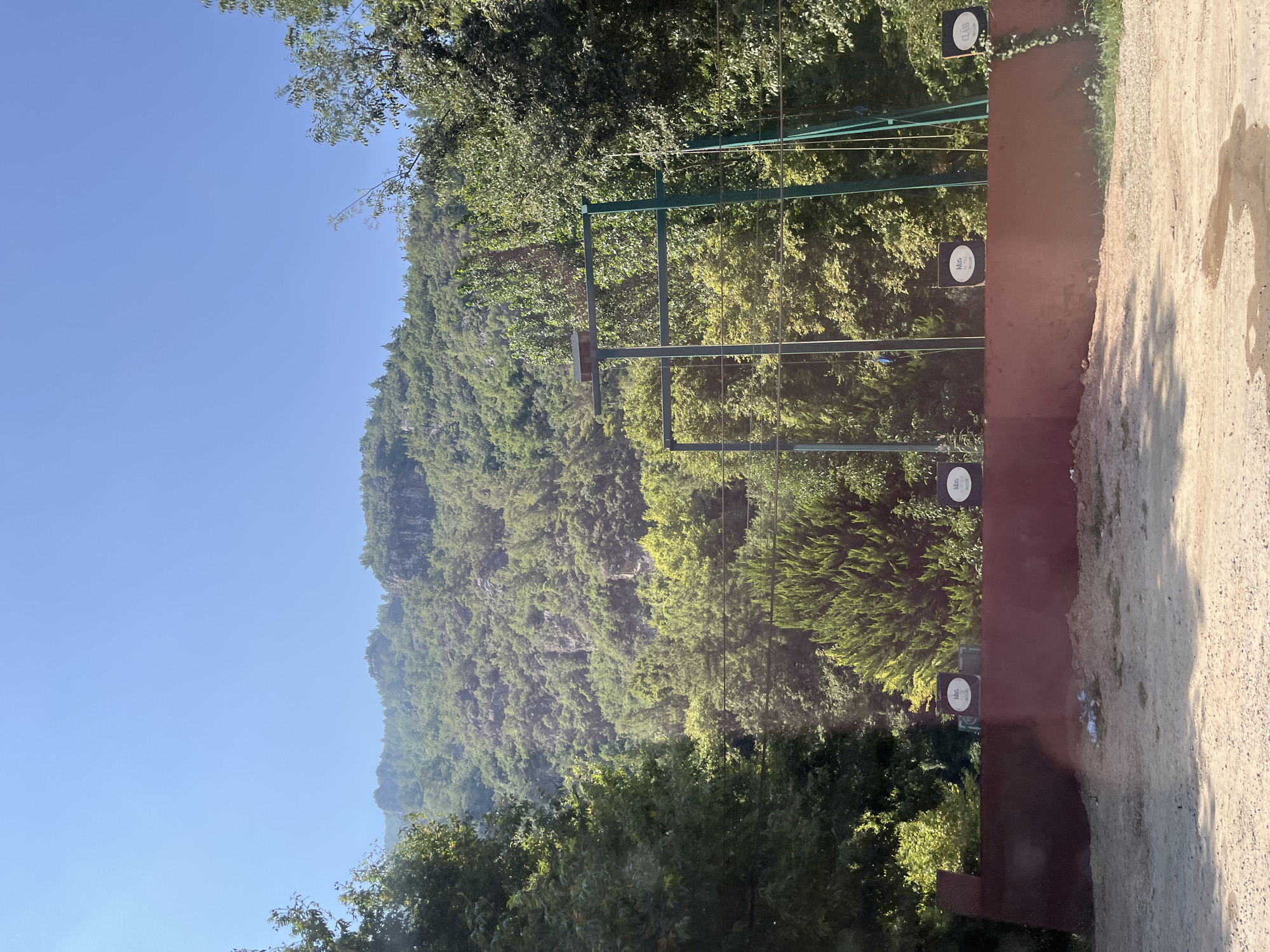 The image depicts a sun-drenched outdoor scene with a focus on a rustic, unpaved area in the foreground, backed by a low, reddish-brown retaining wall. Three circular targets with text and logos are affixed to this wall. Above the wall, a network of green metal poles and wires suggests an athletic or recreational setup, possibly a sports facility. Behind this structure, dense green foliage forms a lush backdrop, with trees of varying types and heights. In the distance, a rugged, tree-covered mountain range rises under a clear blue sky. The overall impression is one of a natural, somewhat unmanicured outdoor setting, possibly in a rural or semi-rural area on the outskirts of Alanya, Turkey, given the environment. There are no people visible in the image.
