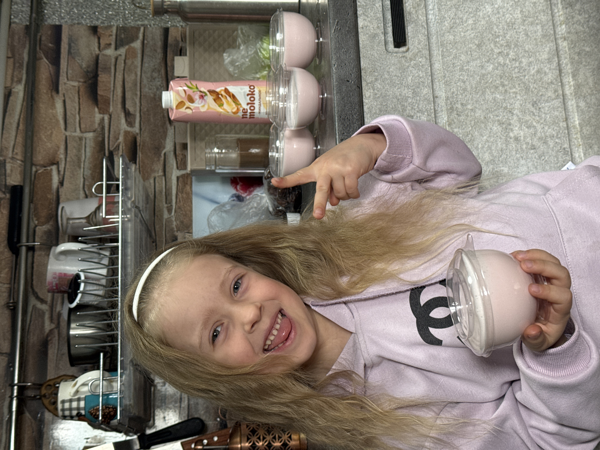 A Caucasian girl with long blonde hair and wearing a lavender sweatshirt with a black logo is holding a sealed cup of pink dessert and pointing at similar desserts in a cupboard. The background includes a carton of milk and kitchenware. The location is in Kurganinsk, Russia.
