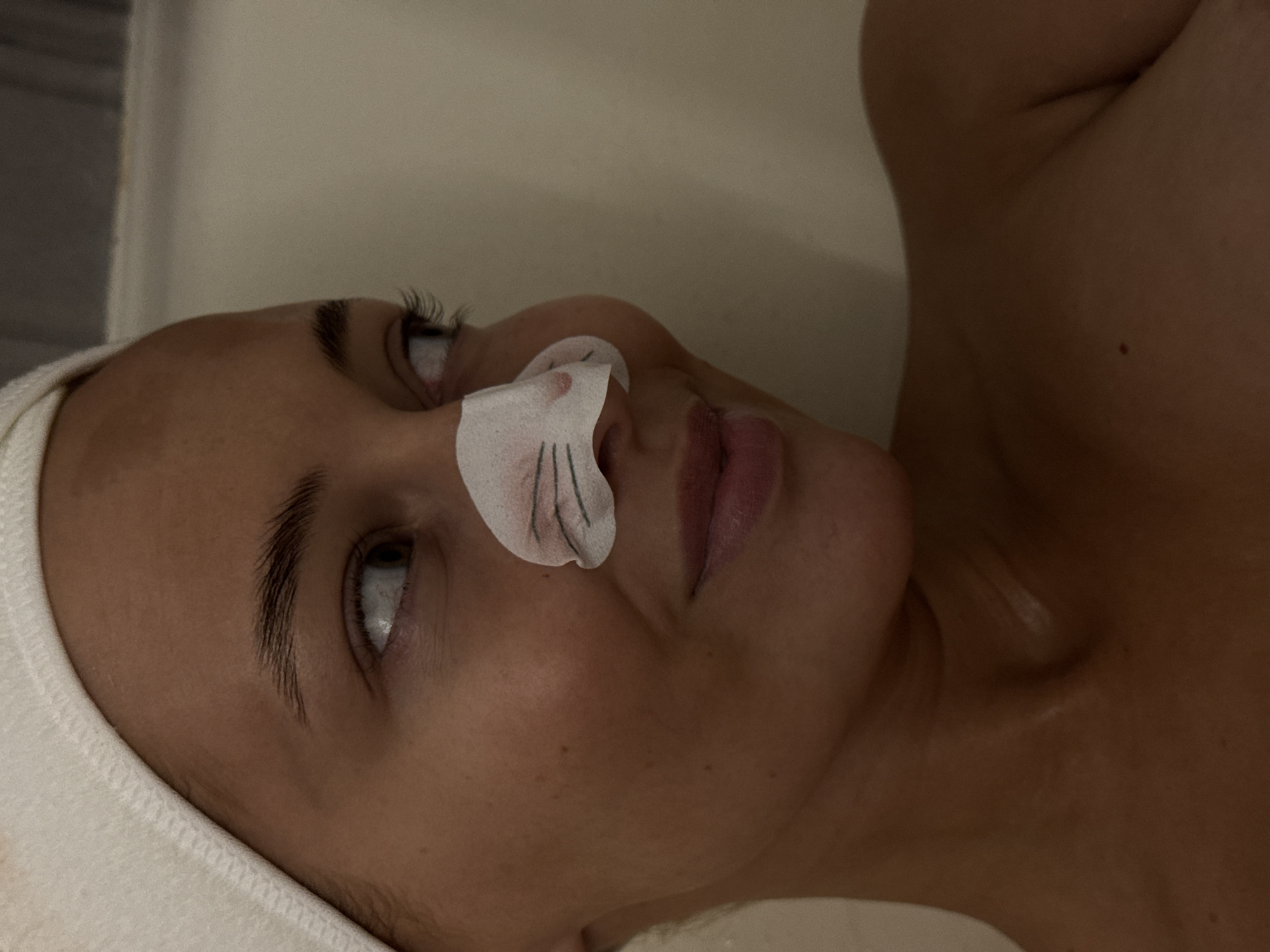 The image shows a fair-skinned person lying down, likely receiving a spa treatment. A white headband is on their head, and a white, cat-shaped strip is on their nose. The person is looking up and to the left. The background is a plain, light color. There is no indication of the location being in Chaikovsky, Russia.