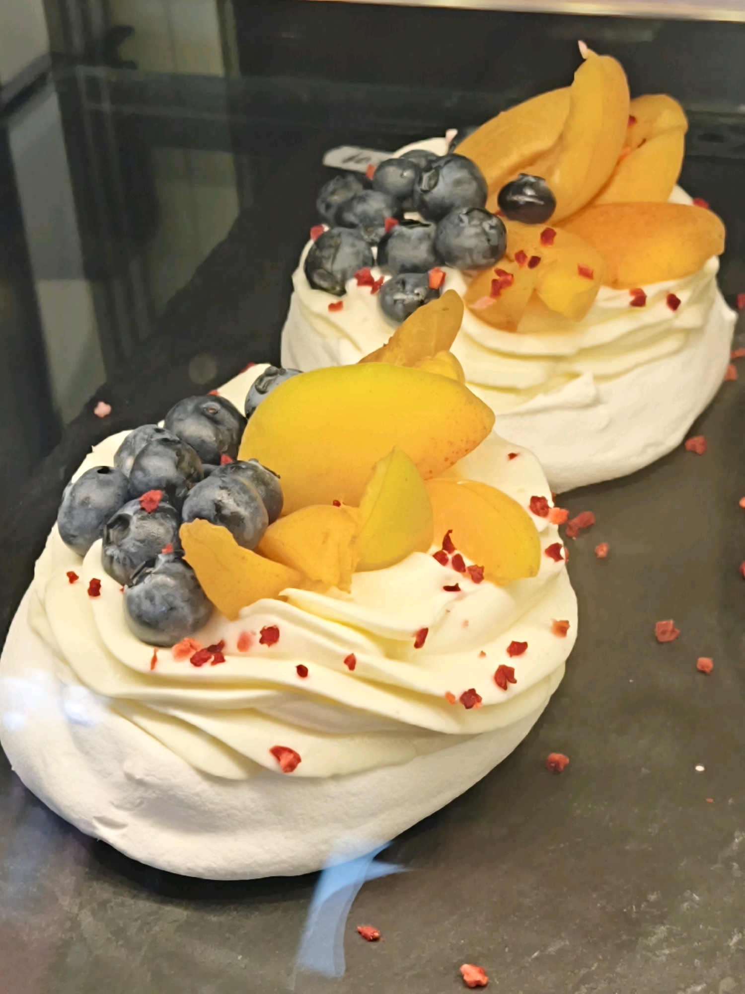The image shows two meringue-based desserts, likely Pavlovas, adorned with fresh fruit and possibly freeze-dried berries. The dessert in the foreground is closer to the viewer and features a swirl of white cream topped with blueberries, slices of apricot, and small red pieces of what appears to be freeze-dried raspberry. The second dessert, slightly further back and to the right, has a similar cream base and is topped with blueberries and apricot slices. Both desserts are presented on a dark, reflective surface. The background is dark and slightly out of focus, suggesting an indoor setting, possibly a cafe or bakery in Tallinn, Estonia, although no specific landmarks are visible to confirm the location.