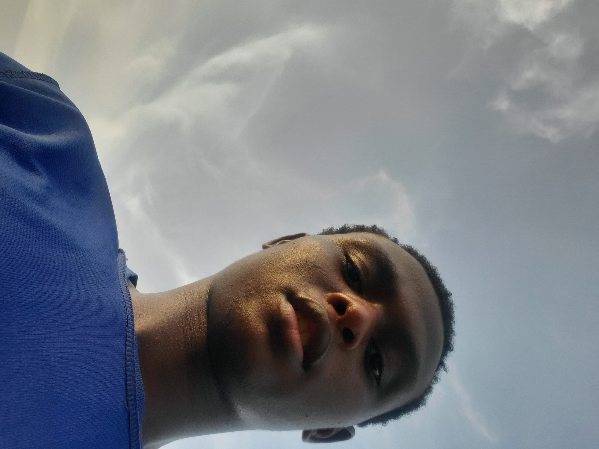 This is a low-angle, waist-up selfie of a young Black man wearing a blue t-shirt. He is looking up at the camera, with a faint smile on his face. The background is a cloudy sky. The image was taken outdoors, likely in Jalingo, Nigeria. The lighting suggests it may be late afternoon or early morning, as the sun is low on the horizon.