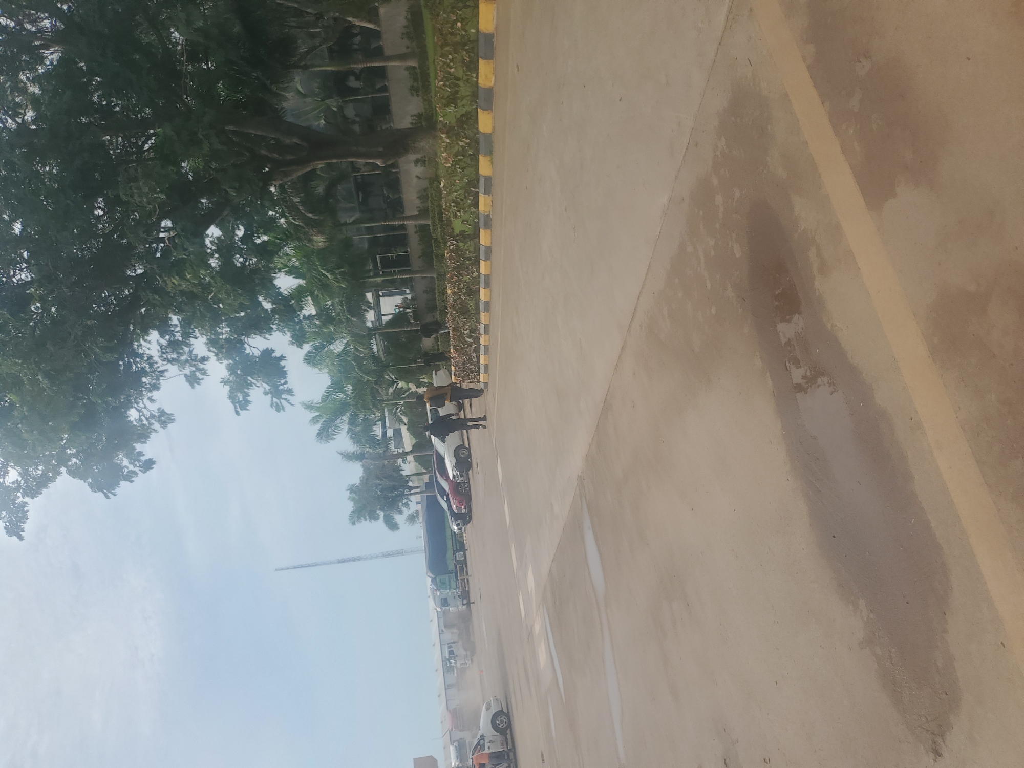 The image captures a scene in Gwagwada, Nigeria, depicting a paved area with several vehicles. In the foreground, the light-colored asphalt shows patches of dampness, suggesting recent rain. A yellow line runs across the pavement, demarcating a parking or traffic lane.
Further back, multiple cars are parked, including a white SUV and a red sedan. Beyond the cars, a fleet of trucks is visible, some of which appear to be cargo trucks with tarpaulin covers. A tall, slender tower, possibly a communication mast, rises in the background against a hazy sky.
The environment is characterized by greenery, with palm trees and other large trees lining the background, interspersed with buildings that appear to be part of a commercial or industrial complex. Two individuals are walking in the mid-ground, facing away from the camera, dressed in casual attire. One is wearing dark clothing, and the other, a lighter brown jacket and pants. Another person, wearing an orange vest, is standing near a white car on the left side of the frame, seemingly engaged in an activity. The overall impression is that of a logistics hub or a large parking area within a business district.