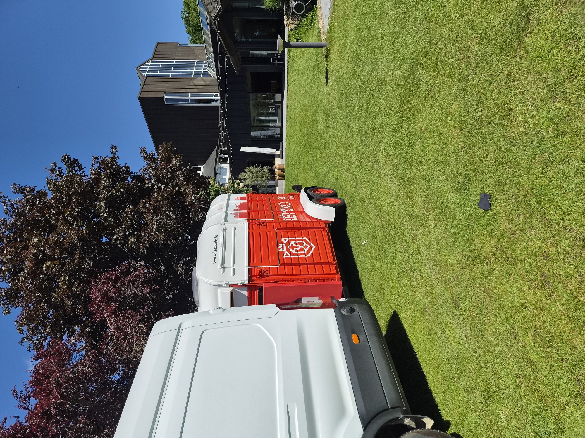 The image depicts a white van connected to a small red and white trailer parked on a green lawn. The trailer has "www.letzkola.lu" printed on its side. In the background, there's a modern, dark-colored house with large windows and an adjacent tree with reddish-brown leaves. The sky is clear and blue. The photograph appears to be taken during daytime with bright sunlight. The location is Lenningen, Luxembourg, indicated by the website printed on the trailer.