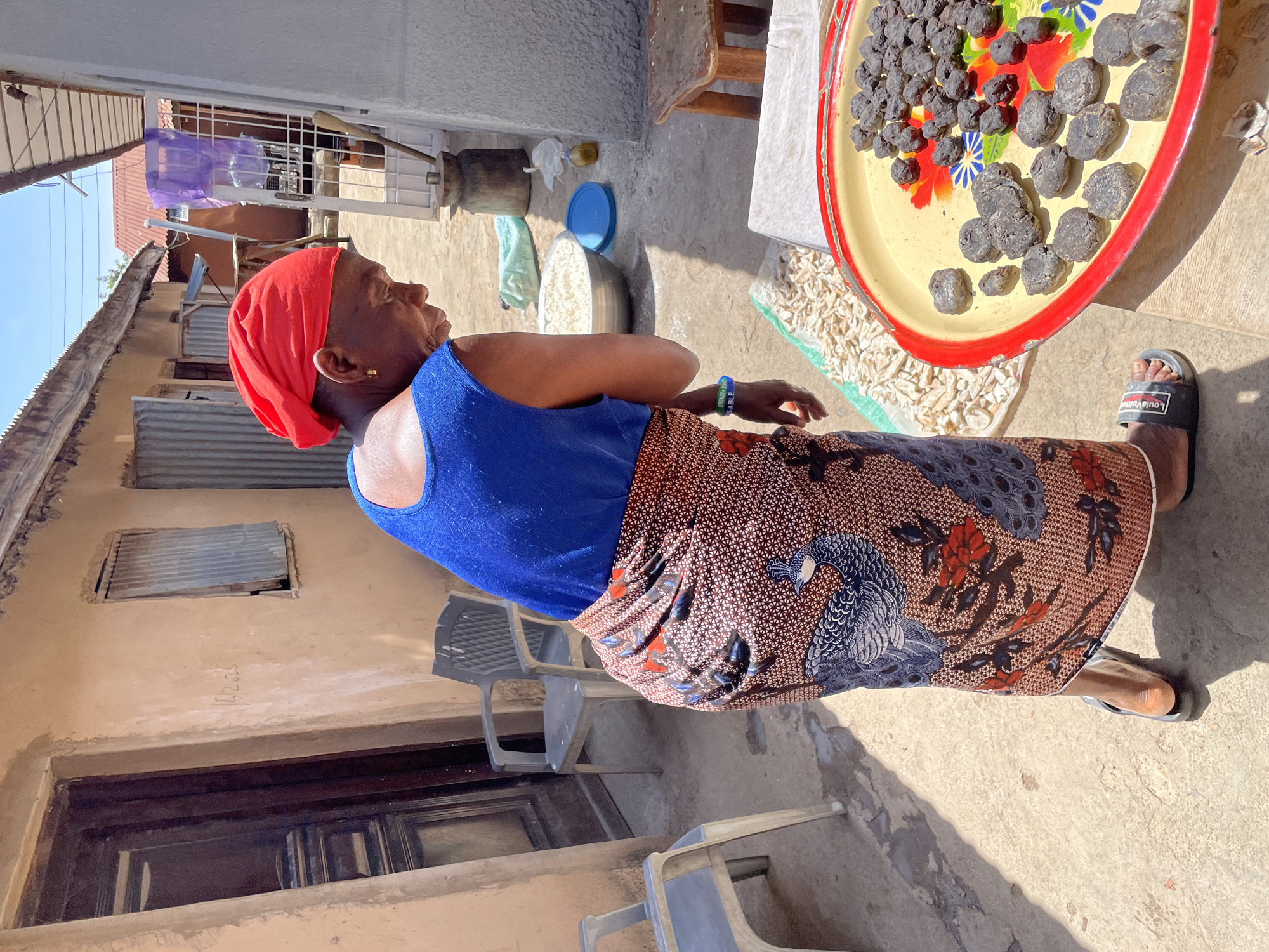 An older woman, seen in profile, stands outdoors in bright sunlight, likely in a courtyard in Jos, Nigeria. She wears a vibrant red headwrap, a royal blue sleeveless top, and a long patterned wrapper featuring peacocks and floral designs in shades of blue, brown, and red. On her left wrist, she has a blue wristband with the text "PRAYER IS ABLE" visible. She wears black "Louis Vuitton" branded slides on her feet. Her gaze is directed slightly upwards and forward, with a neutral expression.
The ground is a mixture of concrete and dirt, illuminated by strong sunlight that creates distinct shadows. To her right, a large yellow metal tray with a red rim holds numerous dark, irregularly shaped, lumpy items, possibly food products like fermented cassava balls, laid out to dry. The tray is decorated with colorful floral patterns. Adjacent to the tray, a pile of white, dried, chipped items, likely cassava or yam pieces, is spread out on the ground, also presumably for sun-drying.
In the background, a silver metal bowl containing a white granular substance, a blue plastic lid, a small yellow bottle, and a light blue cloth are visible on the ground. Several grey plastic chairs and a wooden stool are also present. The surroundings consist of modest buildings with light-colored walls and corrugated iron roofs, typical of residential areas. A window covered with corrugated iron is visible on one wall, which also bears faint vertical writing, appearing to be "42.35." A metal grate in the background has a purple plastic bag and a wooden tool hanging on it. The scene depicts a sunny day, with activities related to food preparation or processing common in the region.