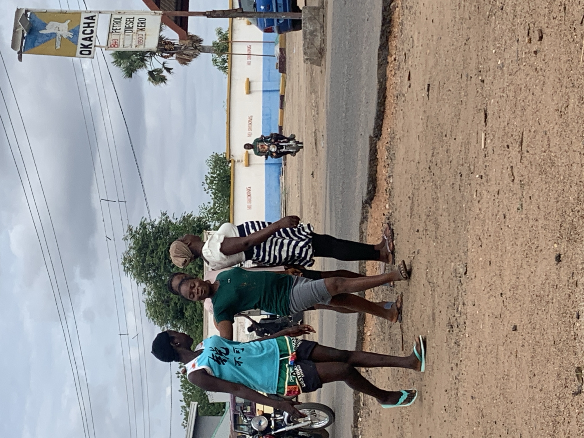 The image captures a street scene in Shendam, Nigeria, under an overcast sky. Three women are walking from left to right on a reddish-brown dirt shoulder alongside a paved road. The woman on the left wears a light blue sleeveless top with dark Chinese characters, dark patterned shorts, and light blue flip-flops. The central woman wears a dark green short-sleeved top, grey shorts, and light brown flip-flops, with her left arm extended towards the first woman's back. The woman on the right wears a white top with puff sleeves, a black and white striped skirt, dark leggings, and dark sandals, with a light brown head covering.
In the mid-ground to the right, a fuel station sign displays "OKACHA" at the top with a blue and yellow emblem featuring a white bird/hand. Below, another section lists "PETROL," "DIESEL," and "KERO" with associated prices, including "840 PETROL" and "650 KERO." Behind the sign, a palm tree and a partially visible blue car are present.
Further in the background, a low white wall with a blue base stripe features multiple red "NO SMOKING" inscriptions. A man on a motorcycle, wearing a dark green shirt, is seen riding in front of this wall. To the far left, another motorcycle is parked. The immediate foreground consists of a rough, stony dirt surface.