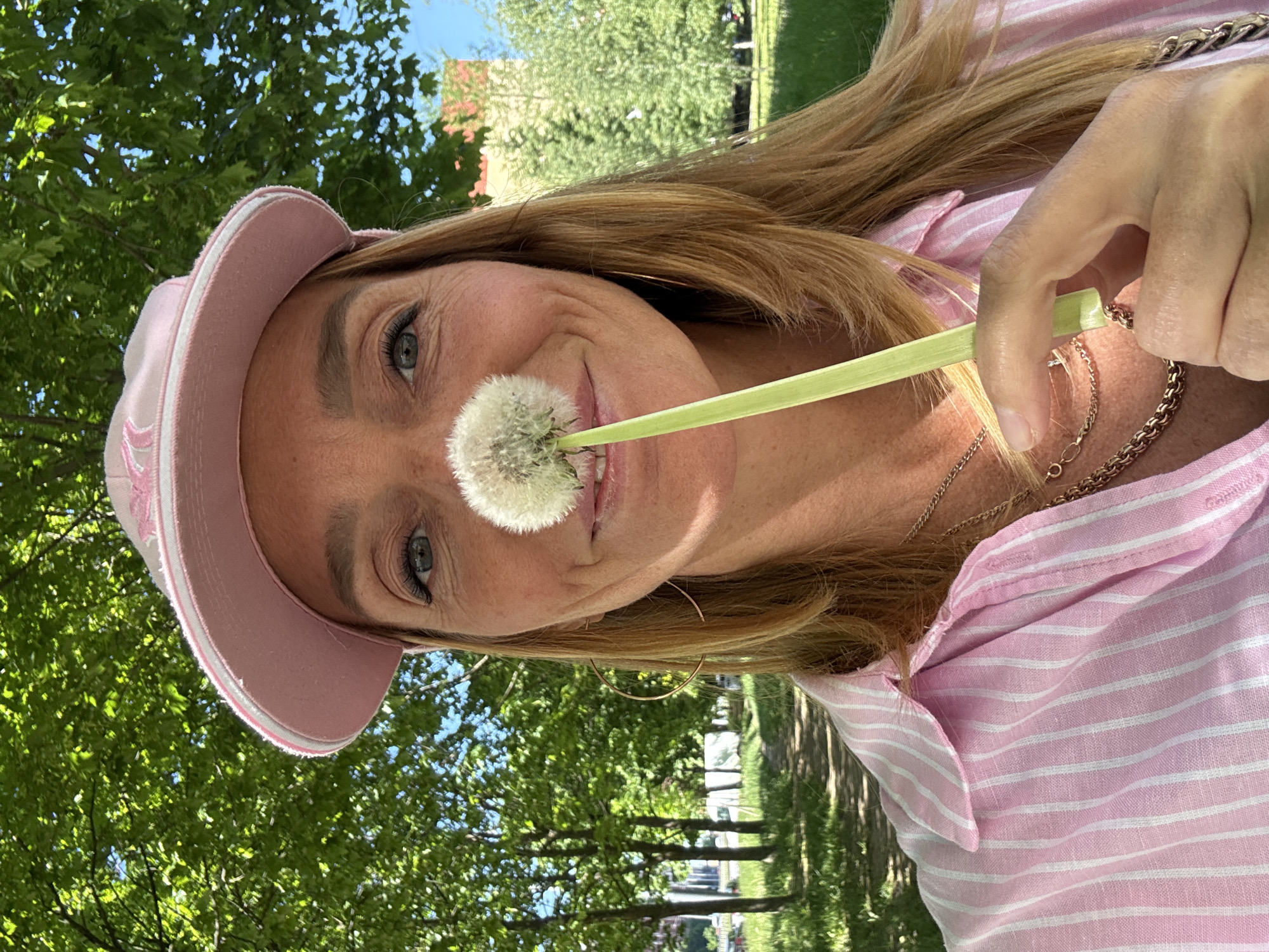 The image features a close-up portrait of a woman outdoors on a bright, sunny day. She is seen from the chest up, smiling gently and looking directly at the viewer. Her long, light brown hair flows over her shoulders, complementing her complexion. She is wearing a pink and white striped shirt and a pink cap or visor, which has an indistinct embroidered logo on the front. A gold-toned necklace is visible around her neck.
The woman is holding a dandelion seed head, commonly known as a puffball, with a long green stem. She delicately positions the fluffy white sphere near her mouth, suggesting she is about to blow its seeds. Her right hand is visible, supporting the stem. Her blue-grey eyes are bright, and her overall expression conveys a cheerful and playful mood.
The background is filled with vibrant green foliage from trees and patches of clear blue sky, indicating pleasant weather and abundant daylight. The setting suggests a park or a natural outdoor area, providing a fresh and natural environment for the portrait. While no specific landmarks are visible in the image, the location context places this scene in Saint Petersburg, Russia. The time of day is clearly daytime, given the bright illumination. No discernible text is clearly visible on the cap or elsewhere.