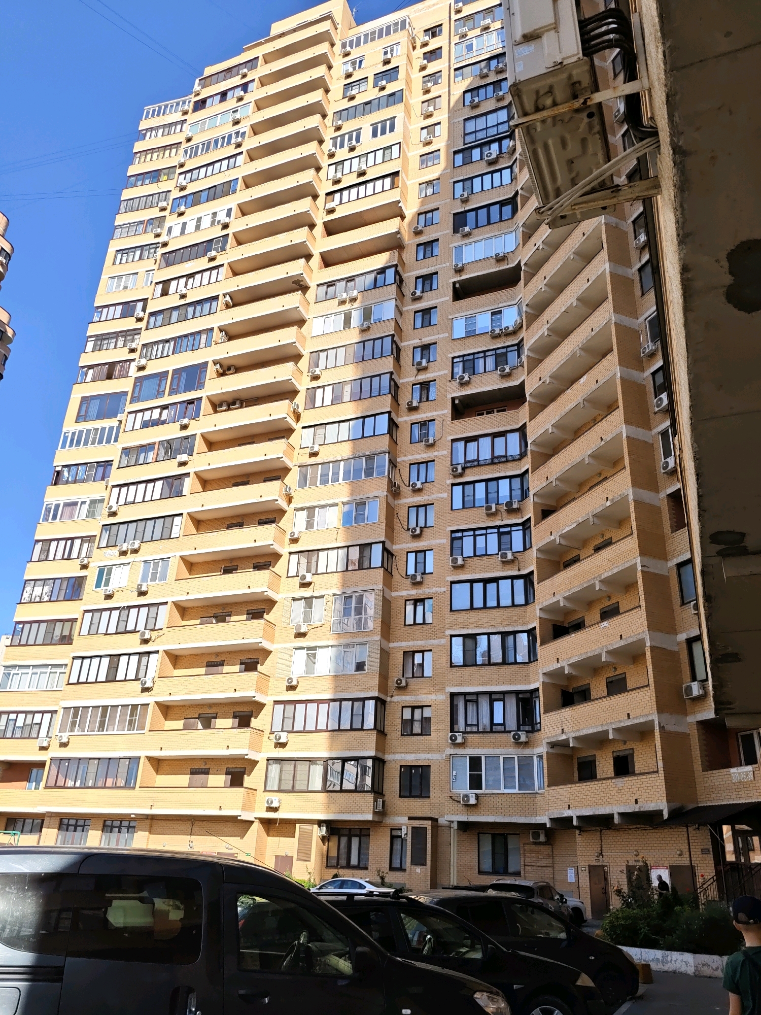 This image captures a high-rise apartment building in Rostov-on-Don, Russia, likely in a residential area. The building is tall, with numerous floors characterized by a light yellow brick facade and beige balconies. Multiple windows, many with dark frames, are visible on each floor. Air conditioning units are mounted outside many of the windows.
In the foreground, the dark, tinted windows and roof of a dark-colored van are prominent, suggesting it is parked in the complex's lot. Another vehicle, a lighter colored car, is partially visible behind the van. The sky above is a clear, bright blue. The overall impression is a typical urban residential scene on a sunny day. The detail "301-05-98" is visible on a sign near a ground-floor entrance, which may indicate a unit number or building identifier.