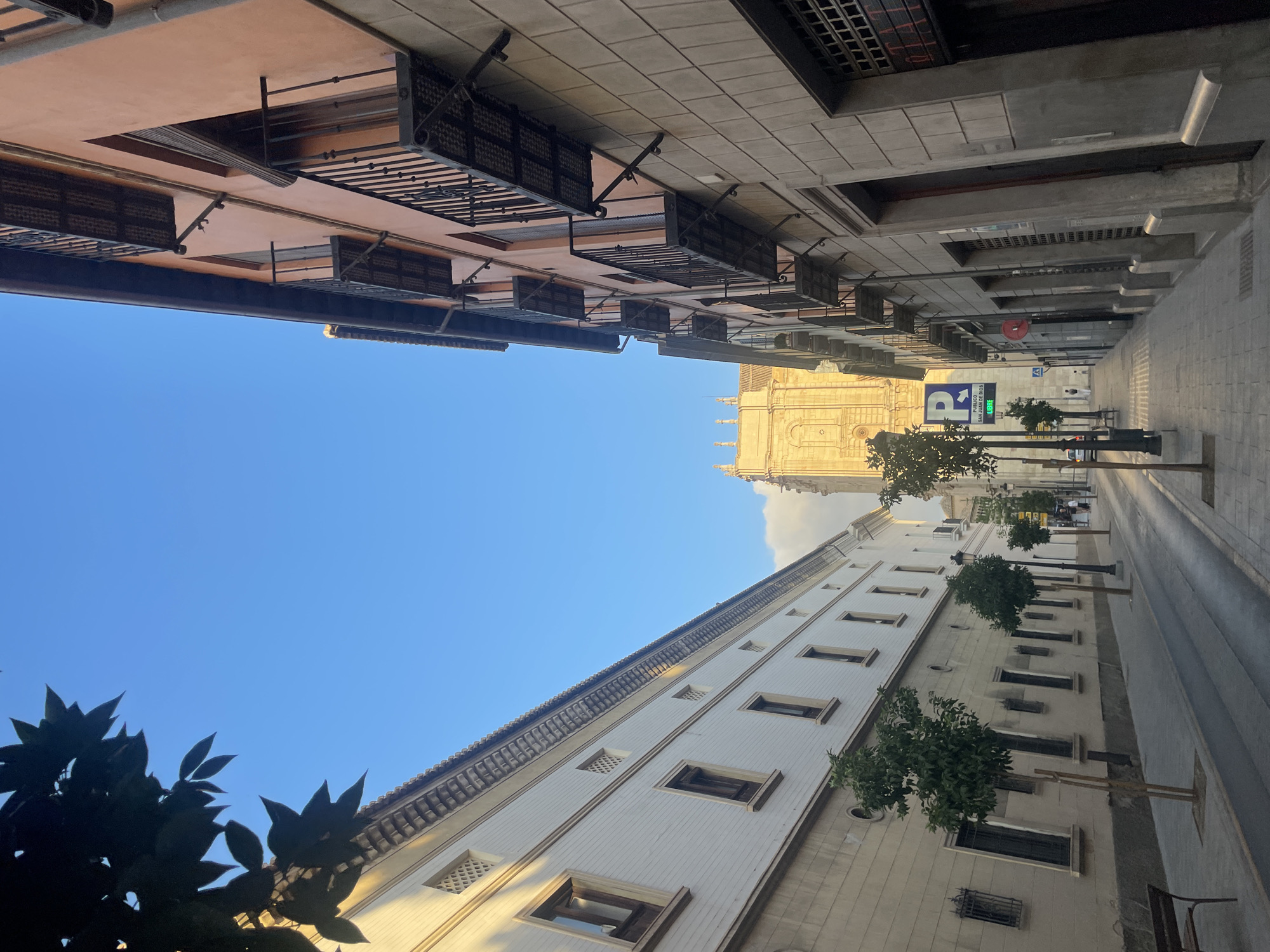 The image depicts a street scene in Granada, Spain, likely in the historic Albaicín or city center, given the architectural style and the presence of a visible parking sign for "San Juan de Dios". The perspective is from street level, looking up a narrow street lined by buildings on both sides.
On the left, a long, white building with multiple windows and decorative friezes runs along the street. It features traditional Spanish architectural elements, including small, dark-paned windows and some openings with decorative lattice work. Small trees are planted along the sidewalk in front of this building.
On the right, a building with an orange-toned facade rises, also featuring multiple windows, some with balconies. These balconies are made of dark metal and are cantilevered out from the building. The lower part of this building is clad in stone blocks.
In the background, a large, ornate building, possibly a church or cathedral, made of light-colored stone and with classical architectural details, is visible. The sky above is a clear, bright blue, with a few wisps of clouds near the horizon. The street itself is paved with cobblestones or similar material. A few streetlights and a bench are also present. The scene is captured during daylight, likely in the late afternoon or early morning, judging by the angle of the light casting shadows. No significant human activity is depicted, with only a few indistinct figures visible in the distance. A green parking sign with a "P" symbol and Spanish text is mounted on a pole in the middle ground.