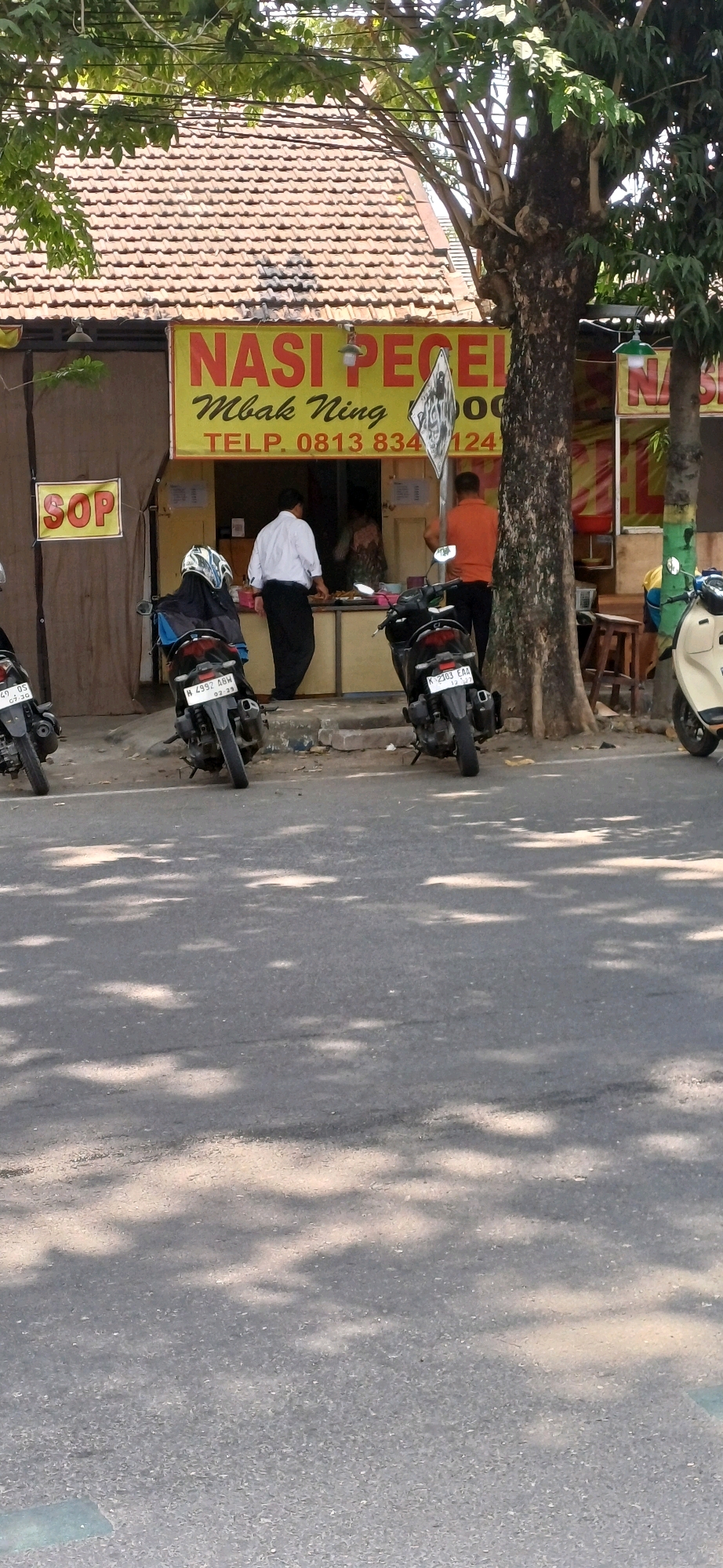 The image depicts the exterior of a small food stall in Kecamatan Pati, Indonesia. The stall has a yellow awning with red lettering that reads "NASI PECEL Mbak Ning" and a phone number "TELP. 0813 834 12". A sign on the left reads "SOP". Two men are visible behind the counter, one in a white shirt and black pants, and another in an orange shirt and black pants. There are several motorcycles parked in front of the stall. The scene is illuminated by sunlight filtering through the leaves of a large tree.