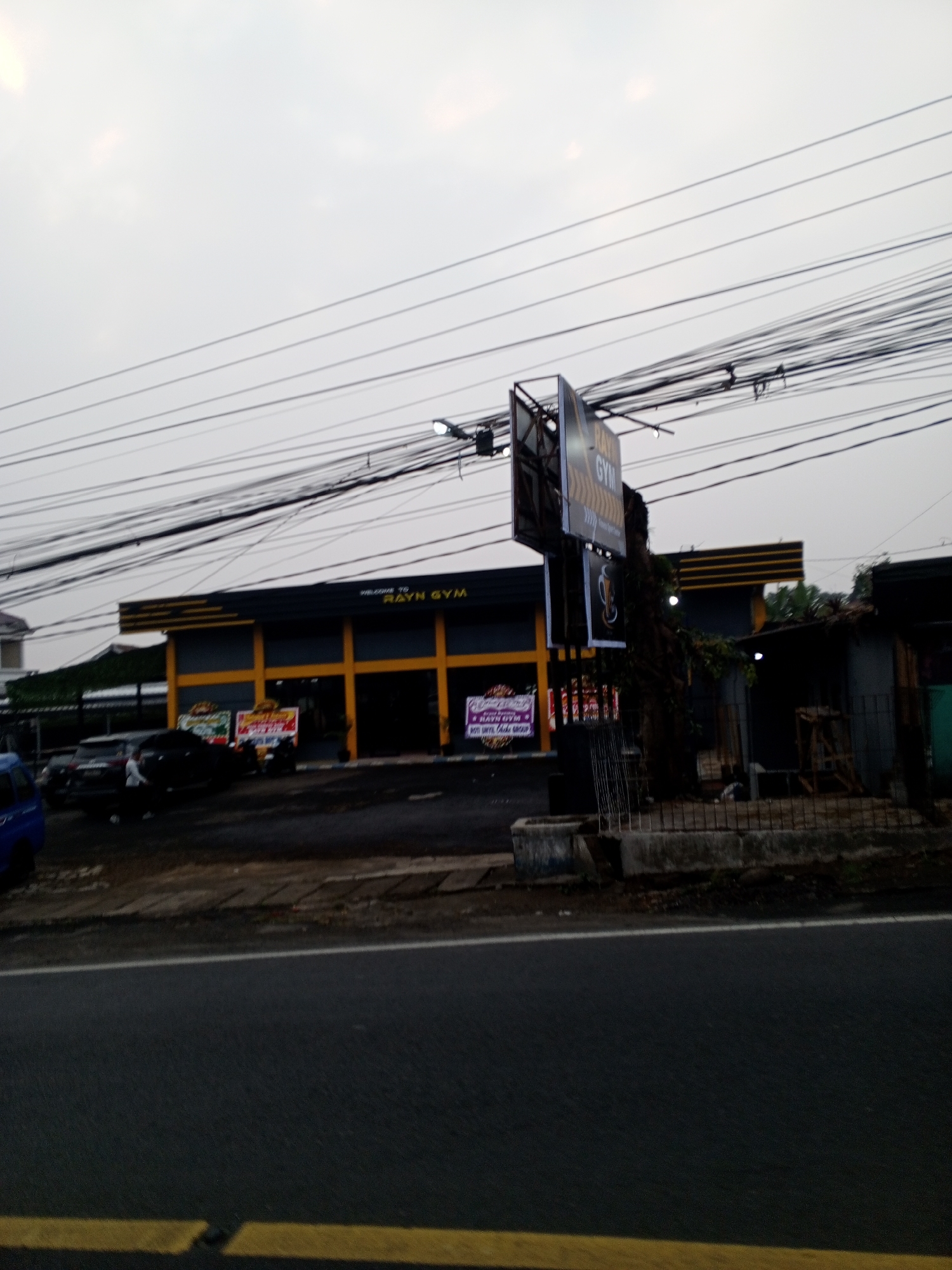 The image shows a building with a sign that reads "RAYN GYM." The building is grey with yellow accents and appears to be a commercial establishment. There are several cars parked in front of the building, and a street runs alongside it. Overhead, a complex network of electrical wires is visible against a hazy sky. The scene suggests an urban or semi-urban environment. There is no information in the image to suggest the location is Dambulla, Sri Lanka.