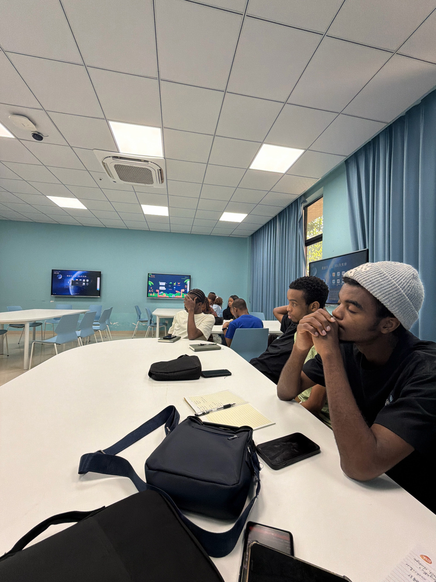 This image captures an indoor scene, likely a classroom or meeting room, with several individuals seated at a large white table. The room has a light blue wall and a white tiled ceiling with recessed lighting. Two large flat-screen televisions are mounted on the wall, displaying content. The foreground features a dark navy messenger bag with a blue strap, a black bag, and a notebook with a pen on the table. One person in the foreground, wearing a light grey beanie and a black t-shirt, is resting their head on their hands, looking down. Another person to their left is wearing a white t-shirt and has their hand covering their face. In the background, more people are visible, also seated. The overall impression is of a learning or collaborative environment. The image is taken from a slightly elevated perspective, looking across the table. The presence of the televisions and the arrangement of the tables and chairs suggest an educational or presentation setting. The specific location of Jiangjin, China is not explicitly indicated in the image itself, but is provided as context.