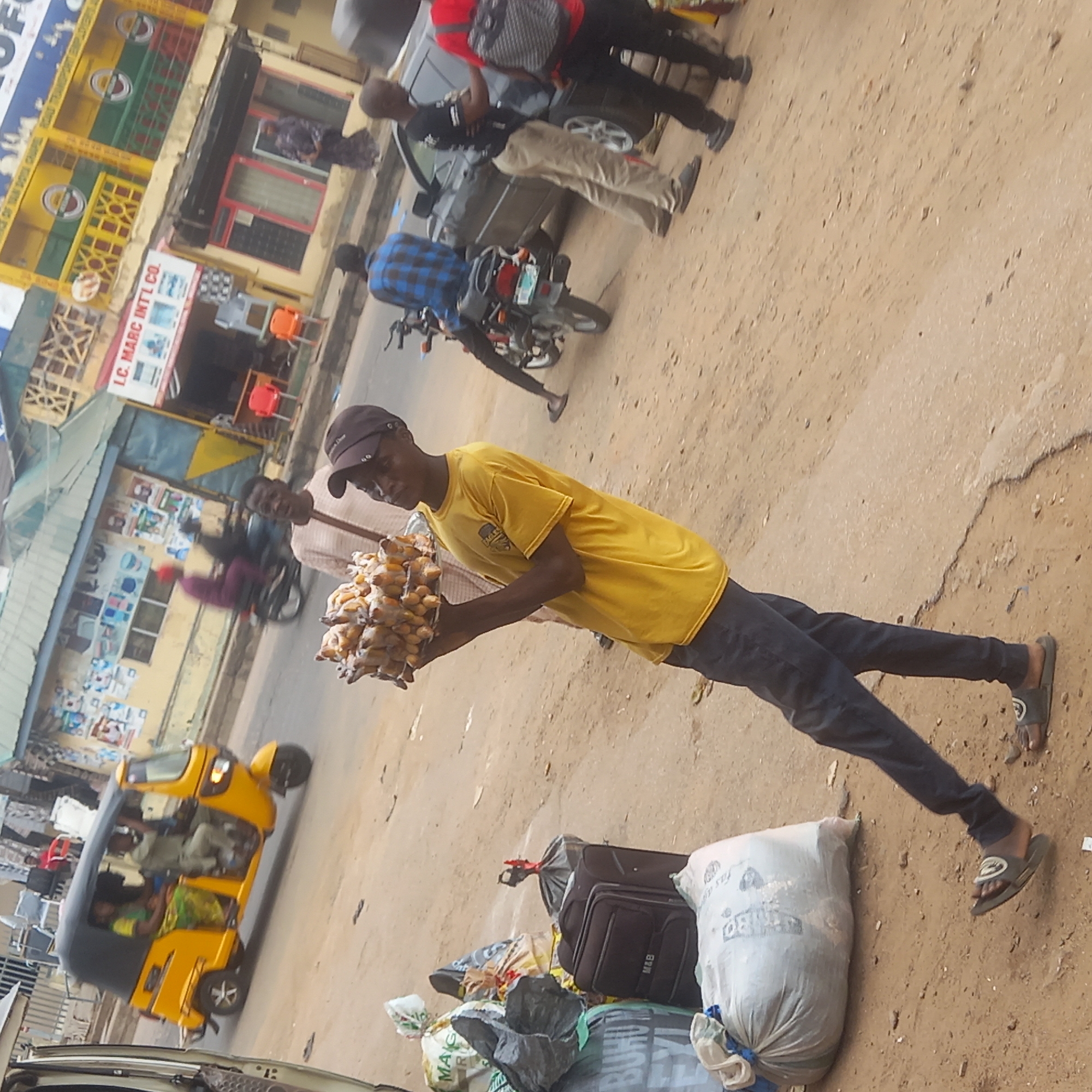 The image shows a man in Sardauna, Nigeria, carrying a tray of what appear to be roasted plantains, a common street food in the area. He is wearing a yellow t-shirt, black pants, and a baseball cap. Behind him is a "Keke Napep," a three-wheeled motorized vehicle used for transportation in Nigeria. Other people are visible in the background, along with parked vehicles and buildings. A sign reads "L.C. MARC INT'L CO." Bags of goods are piled next to the man. The scene appears to be a typical street scene in a busy commercial area of Sardauna.