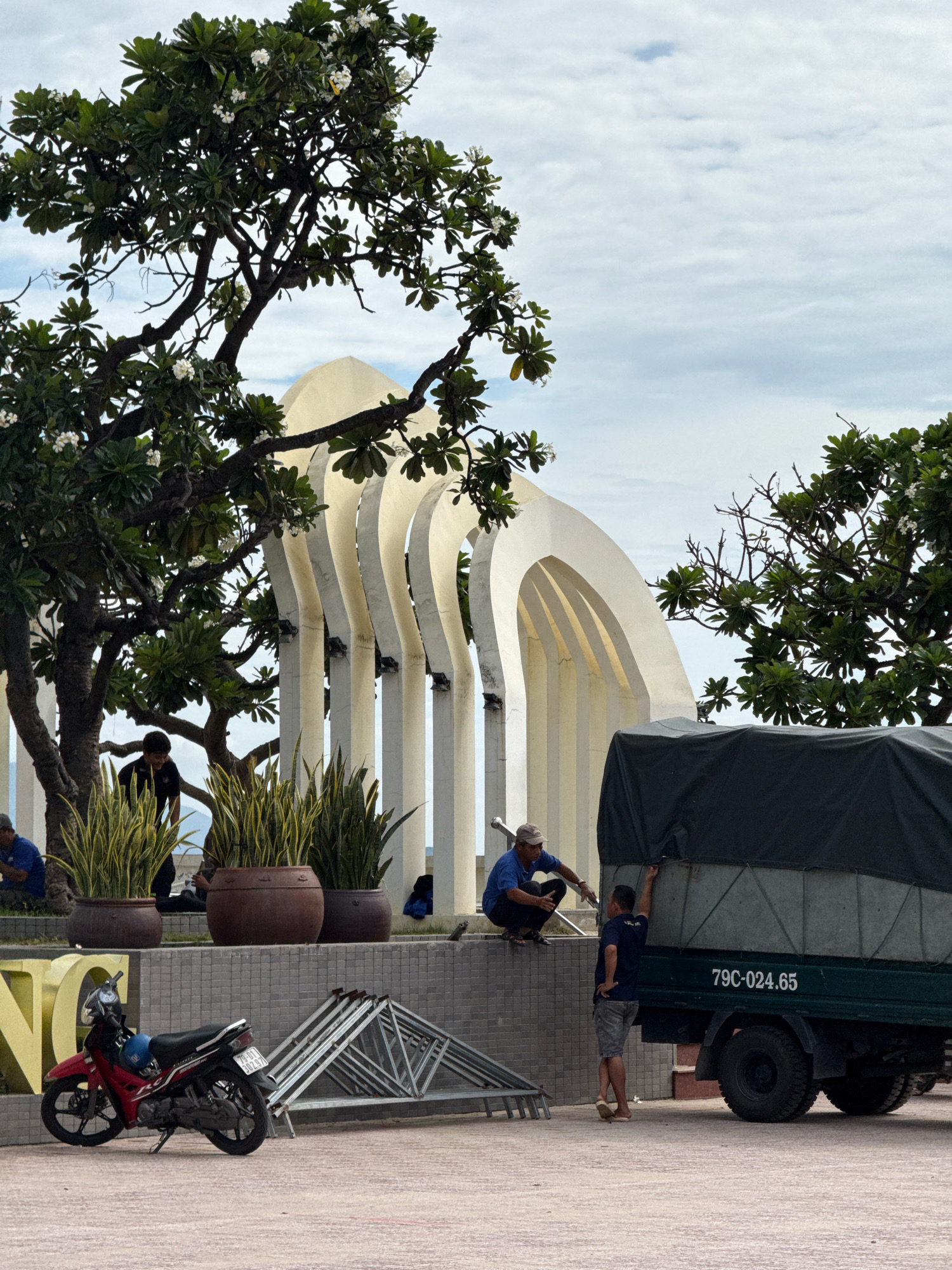 The image depicts an outdoor scene in Nha Trang, Vietnam, likely near a coastal monument or park. In the foreground, a red and black motorcycle is parked to the left of a pile of silver, folded metal scaffolding. To the right, a dark green truck with a black tarp covering its cargo is parked. The truck has a license plate that reads "79C-024.65". Two men are interacting near the truck. One man in a blue shirt and grey shorts is standing with his back to the camera, with his left arm resting on the truck. Another man in a blue shirt and dark pants is crouched down on a low wall, facing the truck. Behind them, a series of white, arched structures are visible, partially obscured by lush green trees with white flowers. A few potted plants are placed on a grey, tiled wall in front of the arches. In the background, the sky is overcast with scattered clouds.
