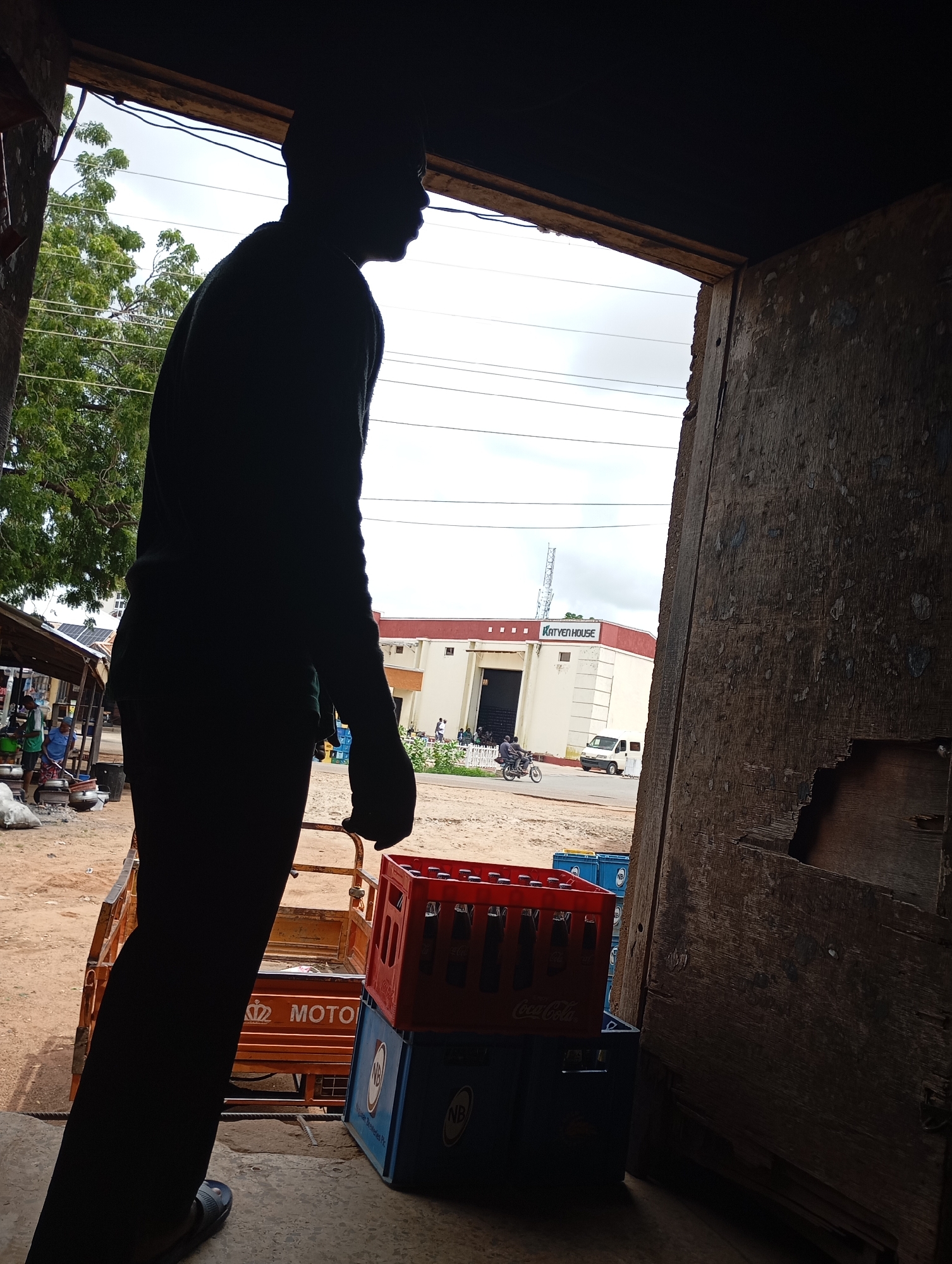 A silhouette of a person stands in the foreground, facing away from the camera and towards an open doorway. The person appears to be male, wearing a dark long-sleeved shirt and dark pants, with dark sandals on his feet. He is holding onto the handle of a three-wheeled motorized cart, which is loaded with crates of beverages. One crate, red and labeled "Coca-Cola," is clearly visible, stacked on top of a blue crate. The cart also has "MOTO" written on its side.
The scene takes place outside, with a street visible beyond the doorway. A building with the words "KATVEN HOUSE" on its facade is prominent in the middle ground. A white van and a motorcycle with two riders are on the street in front of the building. The ground is unpaved and appears sandy. In the background, trees and utility lines are visible against a cloudy sky. The doorway itself is rustic, with worn wooden framing. The overall impression is of a commercial area in Shendam, Nigeria, possibly a shop or distribution point.