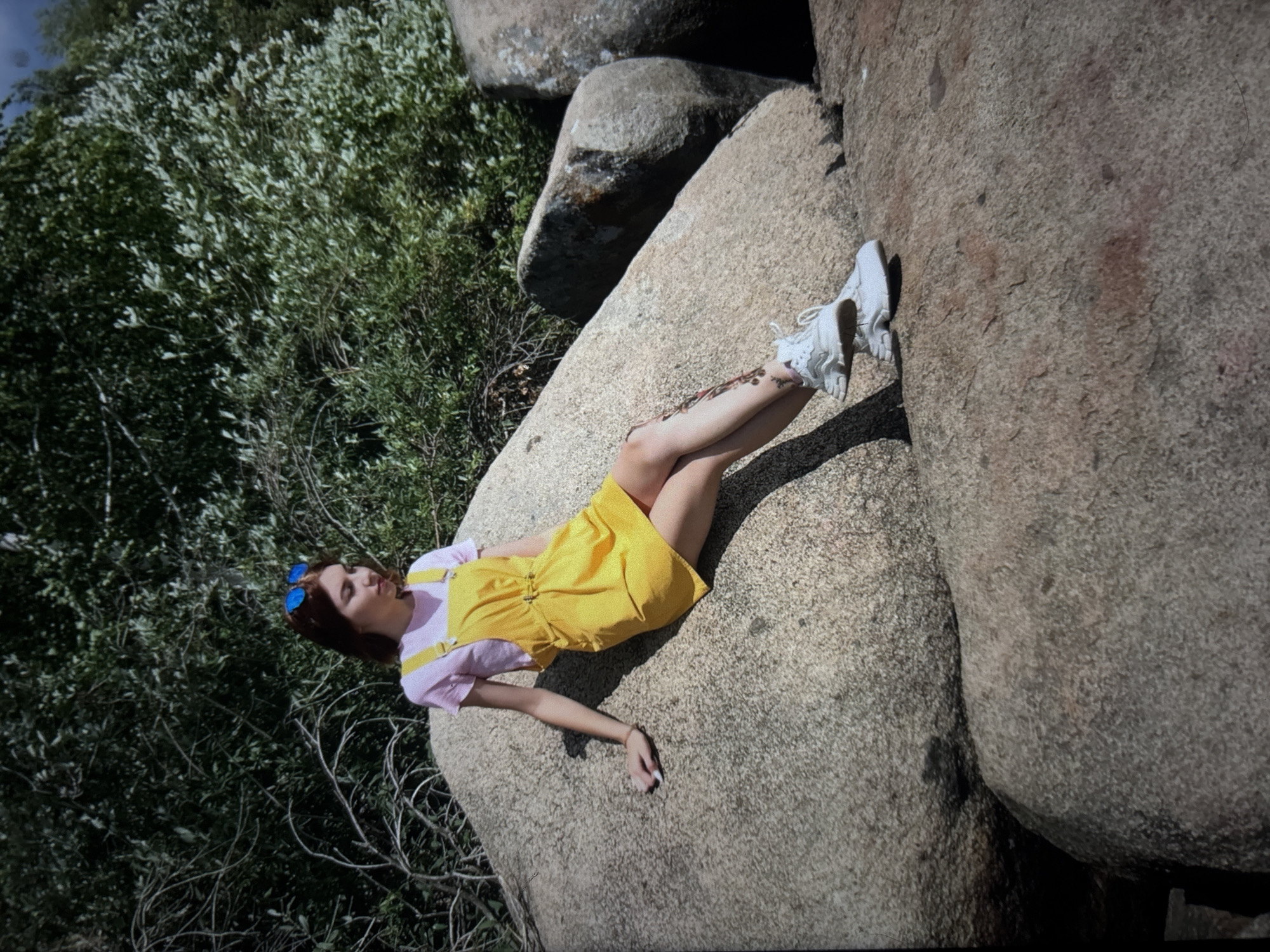 A young woman with auburn hair and blue-tinted sunglasses sits on a large, weathered boulder. She is wearing a pink t-shirt and a bright yellow pinafore dress. Her right arm rests on the boulder, and her left leg is bent with her foot on another rock. She is wearing white athletic shoes. The background consists of lush green foliage and more large rock formations. The image appears to be taken outdoors on a sunny day. The specific location in Omsk, Russia, is not discernible from the visual information provided.