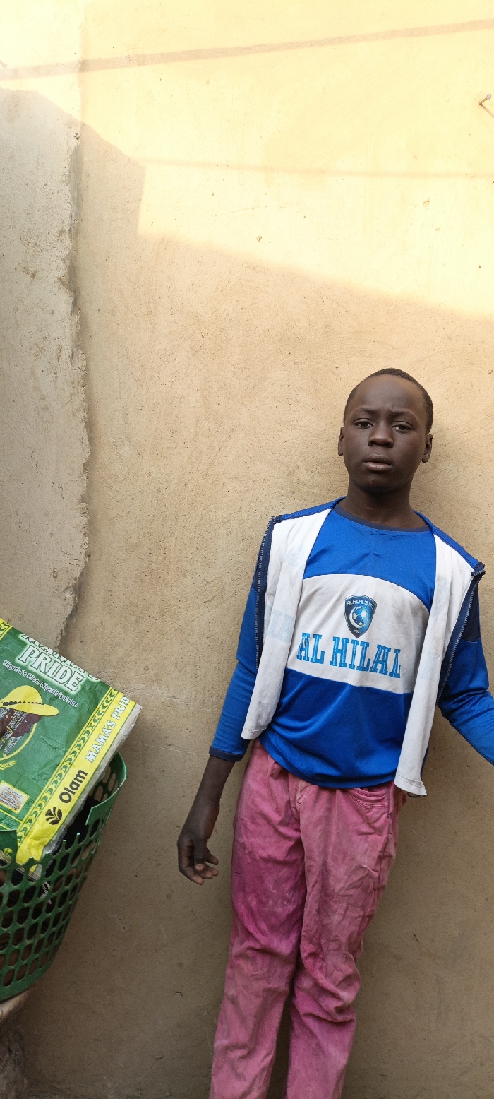 A young male, appearing to be a child or early teenager, stands centrally against a light tan, textured wall outdoors. He has dark skin and looks directly at the camera with a neutral expression.
He is dressed in a blue long-sleeved shirt featuring a white central panel. On this panel, the word "AL HILAL" is printed in blue capital letters, with a crest above it that includes what appears to be "A.H.A.S.C. 1957". Over the shirt, he wears an open, white sleeveless vest or light jacket. His trousers are a faded pink or reddish color and appear worn.
To the left of the boy, partially visible, is a green plastic basket containing a large green and yellow bag. Text on the bag includes "PRIDE", "Nigeria's Rice. Nigeria's Pride", "Olam", and "MAMAS PRIDE".
The scene is brightly illuminated by natural sunlight, casting visible shadows on the wall, suggesting it is daytime with clear, sunny weather. The background wall shows some wear and discoloration. The overall setting is simple and appears to be an outdoor area in Kaura Goje, Nigeria. The boy is not engaged in any specific activity beyond posing for the photograph.