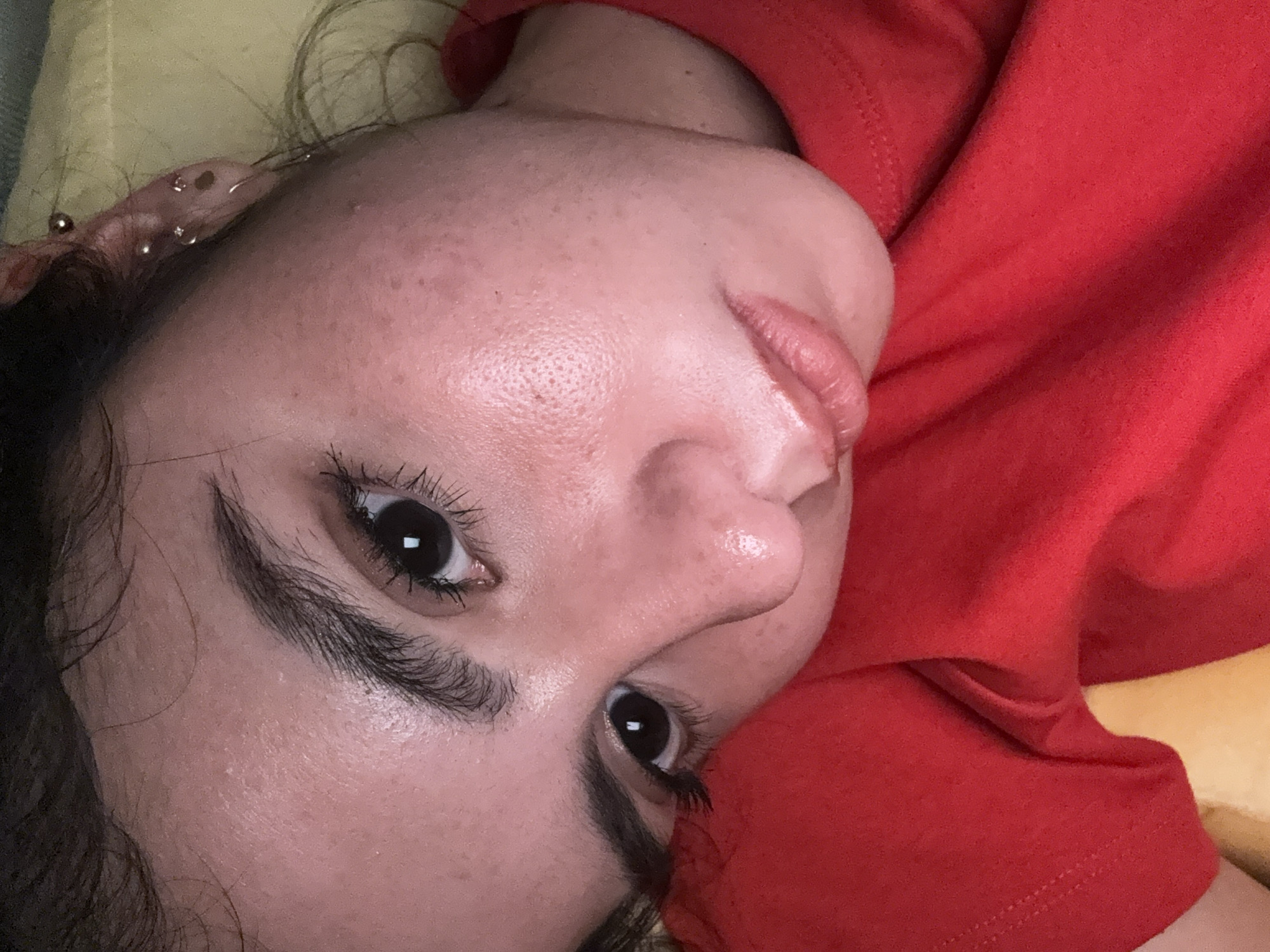 A close-up, rotated image shows a person lying on a yellow surface, possibly a pillow or cushion. The person is wearing a red garment, likely a shirt or top. Their face is prominent, displaying features such as dark eyes with thick eyelashes, dark eyebrows, a nose with visible pores, and slightly glossy lips. The skin shows some texture and minor imperfections. The upper part of an ear with multiple piercings is visible in the top left corner. The background is blurred, further emphasizing the person's face. No specific location in Austin, Texas, can be determined from the visual information provided.