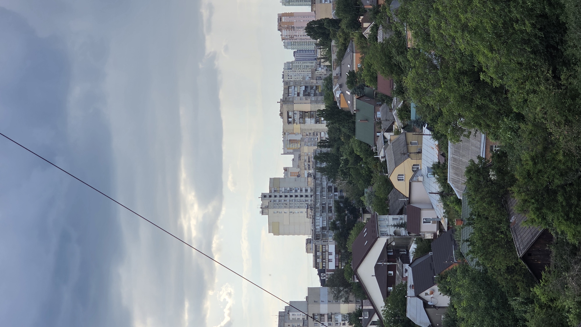 An aerial view of a densely packed residential area in Kyiv, Ukraine, features a mix of multi-story apartment buildings and smaller houses interspersed with lush green trees. The sky is overcast with heavy clouds, creating a diffused light. A thin, dark wire diagonally traverses the upper portion of the frame, running from the top left towards the middle right. The apartment buildings, mostly light-colored, vary in height and architectural style, with many windows visible. The houses below are a mix of styles and colors, with predominantly dark-colored roofs and some with visible chimneys. The foliage is dense and green, suggesting a summer season. There are no people or vehicles immediately apparent in the scene, and the overall impression is one of a typical urban residential neighborhood under a cloudy sky.
