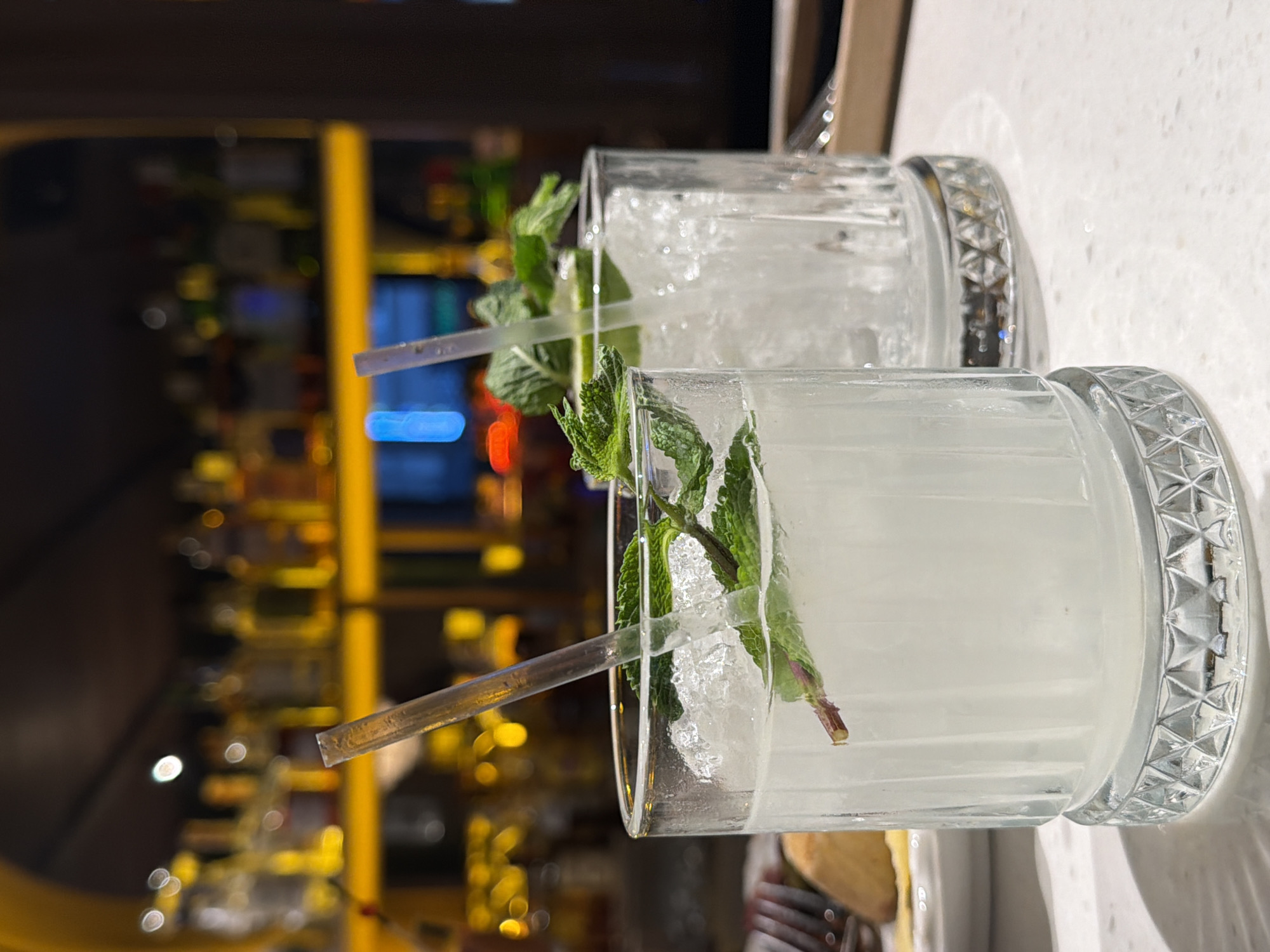 Two glasses of a clear, pale liquid with ice and mint sprigs are the primary subjects. The glasses are made of clear glass with a decorative patterned band around the rim, and have thick glass bases. Each glass contains a clear drink with a substantial amount of crushed ice and a sprig of fresh mint. A clear plastic straw is visible in each glass, partially submerged in the ice and liquid. The background is blurred, showing a dimly lit interior with shelves of bottles, suggesting a bar or restaurant. The lighting is warm and creates bokeh effects with distant lights. The overall impression is of a refreshing alcoholic beverage, likely served in a bar. No people are visible. The scene appears to be indoors, and the time of day cannot be determined. There is no visible text in the image.