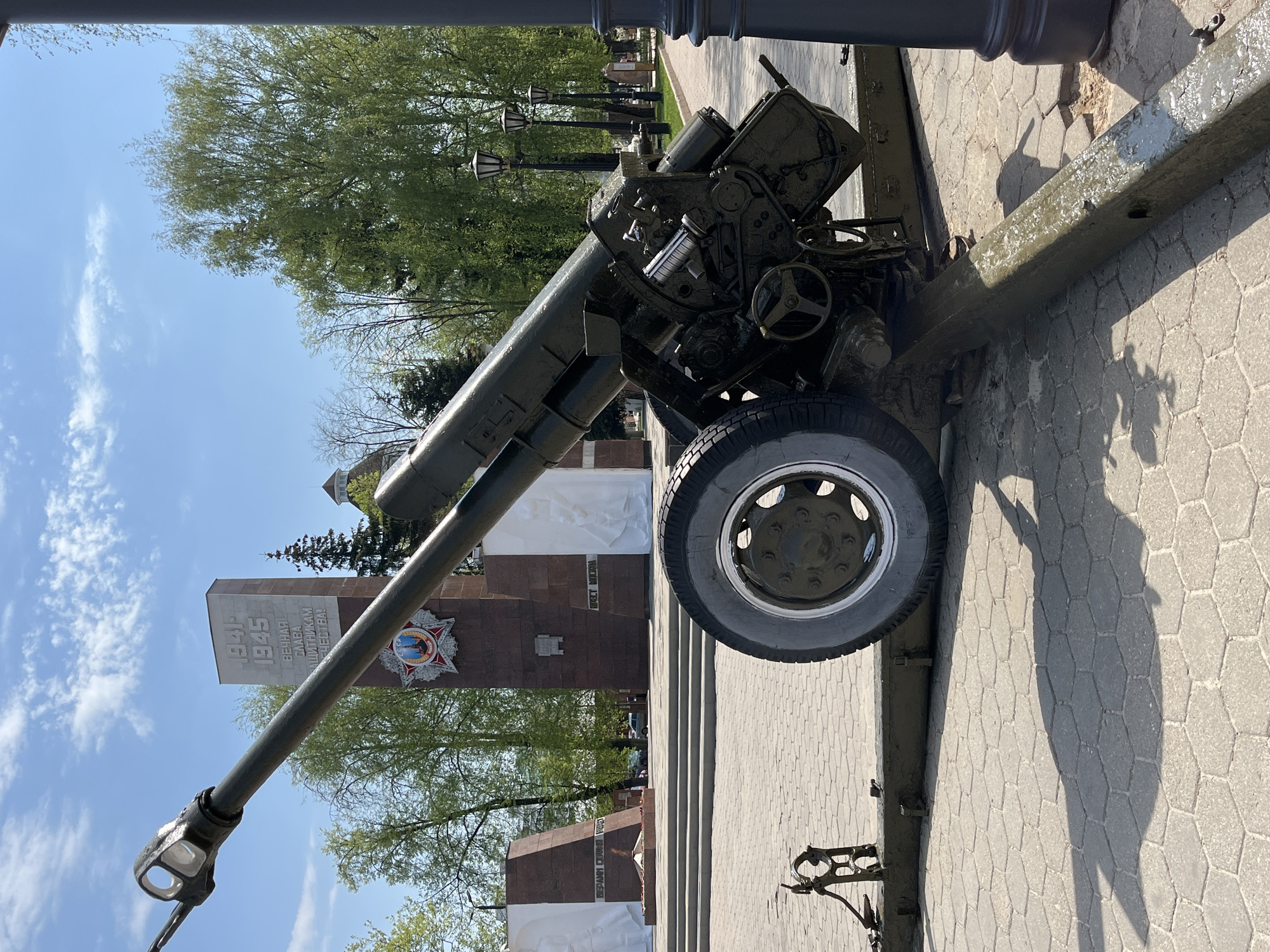 The image shows a Soviet anti-tank gun of World War II vintage, specifically a ZiS-3, on display outdoors. In the background is a memorial complex related to WWII, likely the "Eternal Flame" memorial in the city of Noginsk, Russia. The ZiS-3 is painted in a green color, and its muzzle is pointed towards the sky. The memorial consists of brown and white monuments, with text in Russian including the years "1941-1945" and the phrase "Eternal Glory to the Defenders of the Fatherland." There are trees with green leaves visible in the background and a blue sky with some clouds. The ground is paved with hexagon-shaped stones.