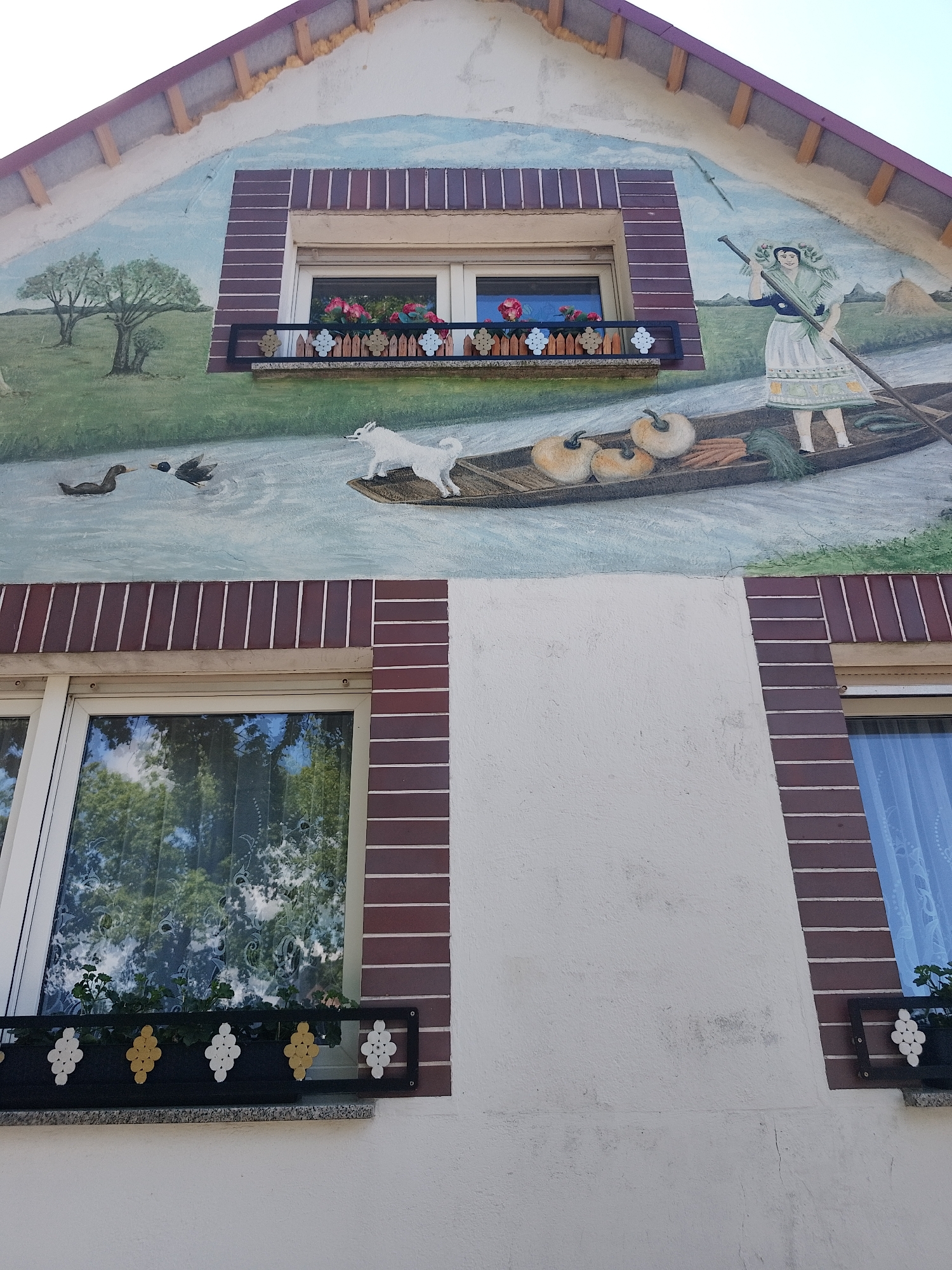 The image shows the exterior of a building, likely a house, in Unknown, Unknown. The top portion of the building features a painted mural, possibly depicting a folk tale or local tradition. The mural shows a woman in traditional clothing standing in a boat filled with produce. A white dog is also in the boat, while two ducks swim in the water nearby. The landscape includes trees and a field.
Below the mural are two windows, each with a flower box. The left window has a clear reflection of trees and sky. The right window has blue curtains partially visible. The building's facade is mostly white with brick-like accents framing the windows.