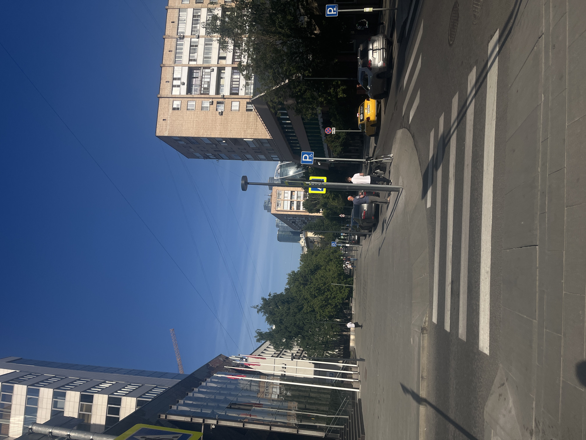 This outdoor photograph captures a bright, sunny urban street scene in Moscow, Russia, under a clear blue sky. The image, seemingly rotated, depicts a street with buildings, vehicles, and pedestrians.
If oriented correctly, the street runs from the foreground into the background, with sidewalks flanking both sides. On the right, a prominent multi-story building with beige brick-like facade and numerous windows stands tall. On the left, a modern building with a reflective glass exterior is visible, and a construction crane can be seen in the distance against the sky.
The road features a clearly marked zebra crossing with wide white stripes. Long shadows are cast across the street and sidewalk, indicating bright daylight, likely mid-morning or afternoon. Several vehicles are present: a yellow taxi cab and a dark sedan are visible on the street, with other cars in the distance.
Along the sidewalk on the right, several street signs are visible: two blue square signs with a white 'P' indicate parking, and a red circular sign with a diagonal line signifies "No Stopping." A yellow square sign with a black pedestrian icon marks the crossing. Several people are seen on the sidewalks; one individual in a white shirt and dark pants stands near a lamppost, while another walks further down. Trees with green foliage line the street, and overhead wires stretch across the vast blue sky. The scene depicts a typical, active city environment.