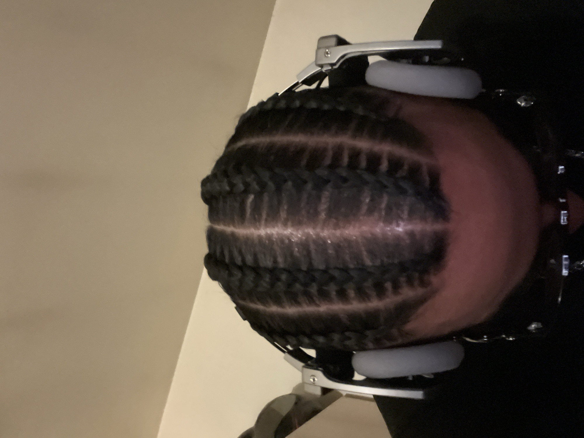 A close-up, angled, indoor shot shows the head of a person with cornrow braids, wearing headphones. The headphones are gray with silver accents. The background consists of a light-colored wall on the left and a dark area on the right. The person's skin is visible between the braids. There is no indication that the location is in Paris, France.