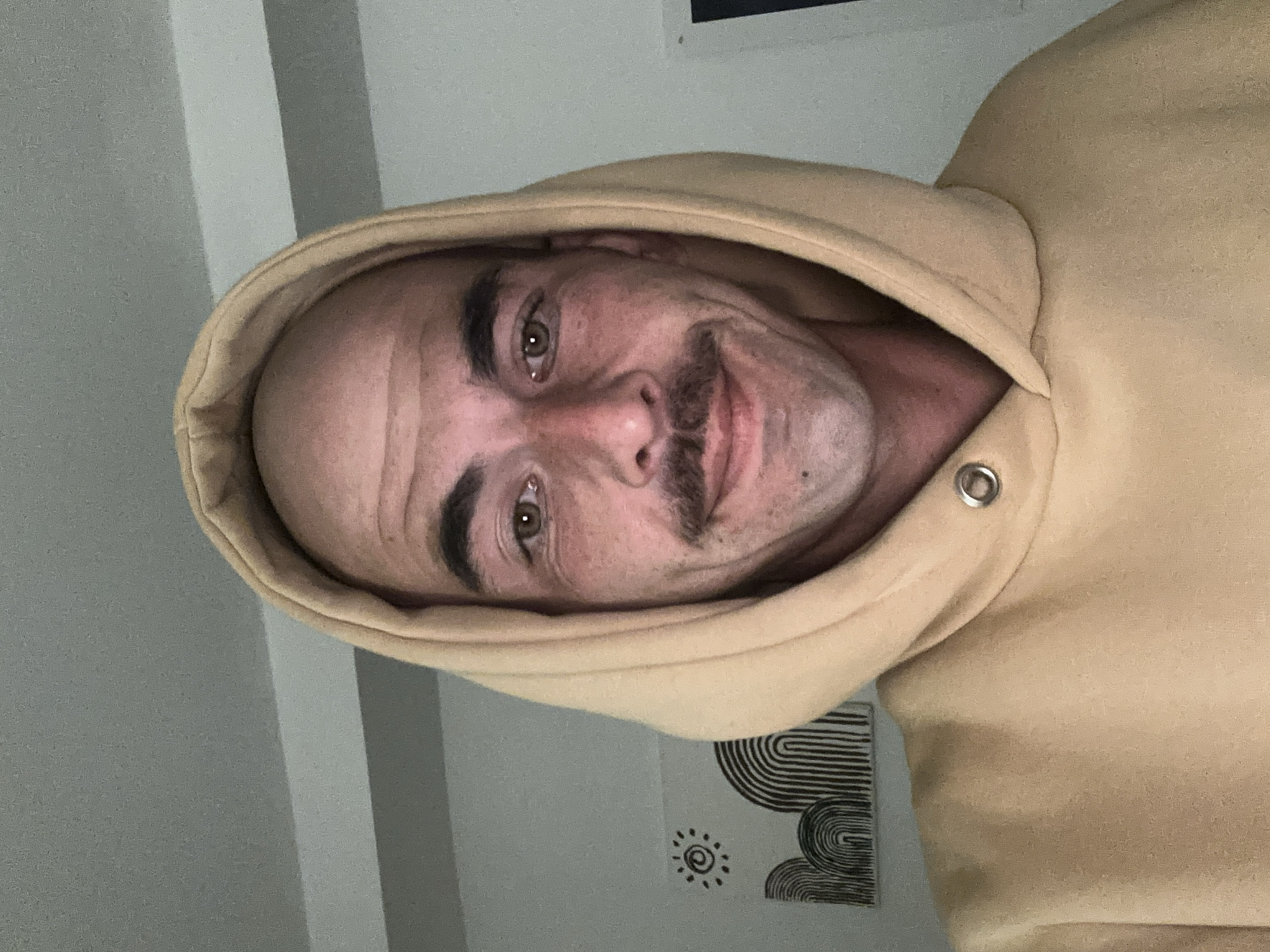 This close-up portrait, captured in Budva, Montenegro, depicts an adult male looking directly at the camera with a slight smile. The subject presents with a bald forehead, prominent dark eyebrows, hazel eyes, and a distinct dark mustache. Freckles are visible on his nose and cheeks, which exhibit a faint reddish hue. He wears a light tan hooded sweatshirt with the hood up, covering the top of his head. A silver-colored metal eyelet is discernible on the hoodie fabric near his right collarbone area. The background consists of a plain, light-colored wall. Mounted on the wall to the left of the subject are two small, square art pieces: the upper one displaying a black stylized sun, and the lower one featuring multiple parallel black arched lines. A faint dark vertical line is visible along the far right edge of the frame. The scene is illuminated by overhead lighting.