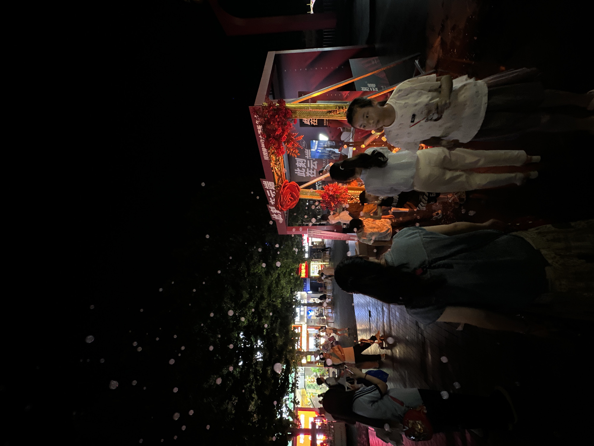 This image captures a nighttime outdoor scene in Chongqing High-tech Zone, China. The environment is primarily lit by artificial sources, casting shadows and highlighting specific areas. Numerous translucent bubbles are scattered throughout the upper and middle portions of the frame, reflecting ambient light.
In the mid-ground, on the right side, a temporary promotional booth or structure is visible. This structure is predominantly red and gold, featuring large, intricate red floral or rose-like decorations. Banners on the booth display Chinese characters, notably "此刻在云端" (Cǐkè zài yúnduān - At this moment in the cloud) and "短视频创作大赛报名" (Duǎn shí pín chuàngzuò dàsài bàomíng - Short video creation contest registration).
Multiple individuals populate the scene. In the immediate foreground, to the right, a young female, estimated to be a pre-teen, with dark hair, wears a white patterned t-shirt and a dark skirt, holding a clear cup with a red straw. To her left and slightly behind, an adult female with long dark hair, viewed from the back, wears a light blue short-sleeved denim-style top and a patterned skirt, walking away from the camera's perspective. Further left, a person in a light blue short-sleeved top and dark pants, carrying a red shoulder bag, is seen from behind, holding a mobile phone horizontally. Near this individual, another person in a white t-shirt and dark pants stands beside a small object with wheels. Several other adults and children are partially visible around and within the vicinity of the promotional booth.
The ground surface is paved, exhibiting reflective properties suggesting dampness, possibly from burst bubbles. In the mid-ground, to the left, several large trees with dense, dark foliage are present, illuminated by background light. Behind the trees, a row of commercial buildings with brightly lit facades, displaying various signs and windows, lines what appears to be a public street or plaza. The sky above is dark and cloudless.