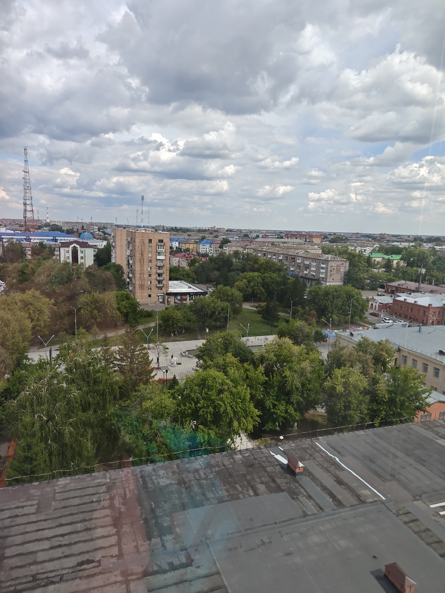 The image depicts a wide, overcast view of a city, likely Petropavl, Kazakhstan. In the foreground, a dark, weathered rooftop with distinct horizontal seams and white markings dominates the lower portion. Beyond the rooftop, a dense canopy of green trees forms a midground layer. Within this verdant expanse, several paved pathways and a small plaza are visible, with two individuals walking. A multi-story brick building with numerous windows is a prominent feature, situated behind the trees. Further into the background, a mix of residential buildings, some with blue roofs, and other structures are scattered across the cityscape. A tall radio tower stands prominently on the left horizon, and a distinct blue dome structure is visible among the buildings. The sky is filled with a dramatic arrangement of grey and white clouds, suggesting an unsettled weather day. The overall impression is of a typical urban environment with a significant presence of green spaces and diverse architectural styles.