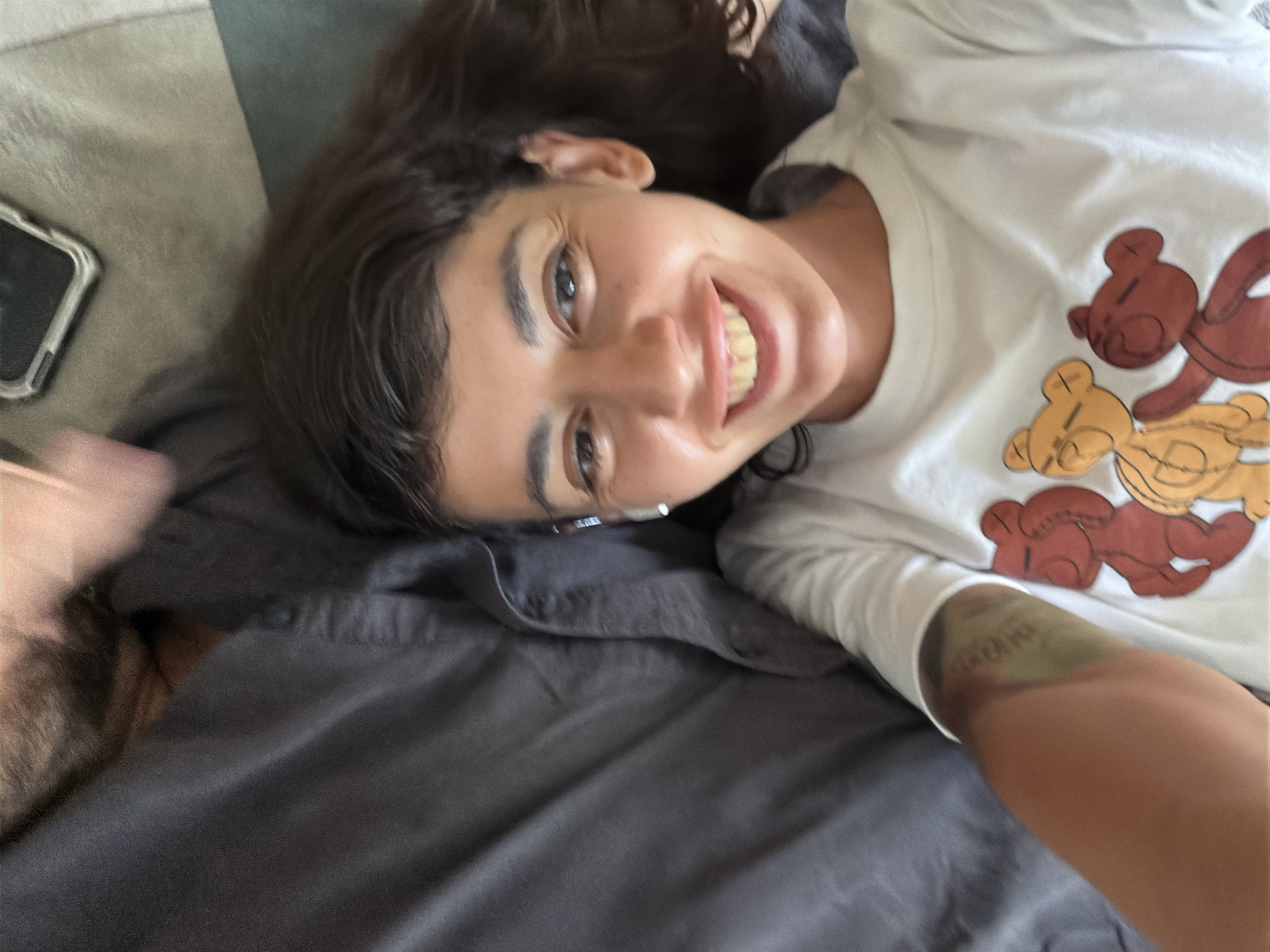 A young woman is lying down on a bed, looking up and smiling at the camera. She is wearing a white t-shirt with a graphic of three teddy bears. She has dark, wavy hair and is wearing white earbuds. Her left arm is visible, with a green bruise and some text tattooed on it. The background is a dark gray duvet and a lighter gray pillow. There is a corner of a phone case visible in the upper right of the frame. The scene appears to be indoors, possibly in a bedroom.