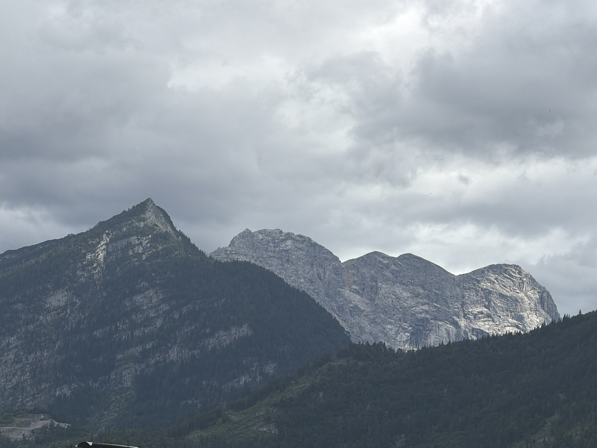 This image captures a scenic view of rugged, tree-covered mountains under a cloudy sky, likely in Torren, Austria. The foreground is dominated by the dark green slopes of forested mountains, with their varied textures and dense foliage. Rising behind these, the stark, grey peaks of higher mountains are visible. These peaks are characterized by their rocky, weathered surfaces, showing distinct geological layers and formations. Some areas of the mountains are illuminated by sunlight breaking through the clouds, creating a contrast with the shaded parts.
The sky is overcast with heavy, grey clouds, suggesting a temperate or cooler climate, possibly late afternoon or an approaching storm. The lighting is diffused, contributing to the somewhat muted color palette of the scene.
There are no people or prominent objects in the frame, focusing solely on the natural landscape. The overall impression is one of natural grandeur and the imposing presence of the alpine environment. The image appears to be a still photograph, capturing a moment of natural beauty. No text is visible within the image.