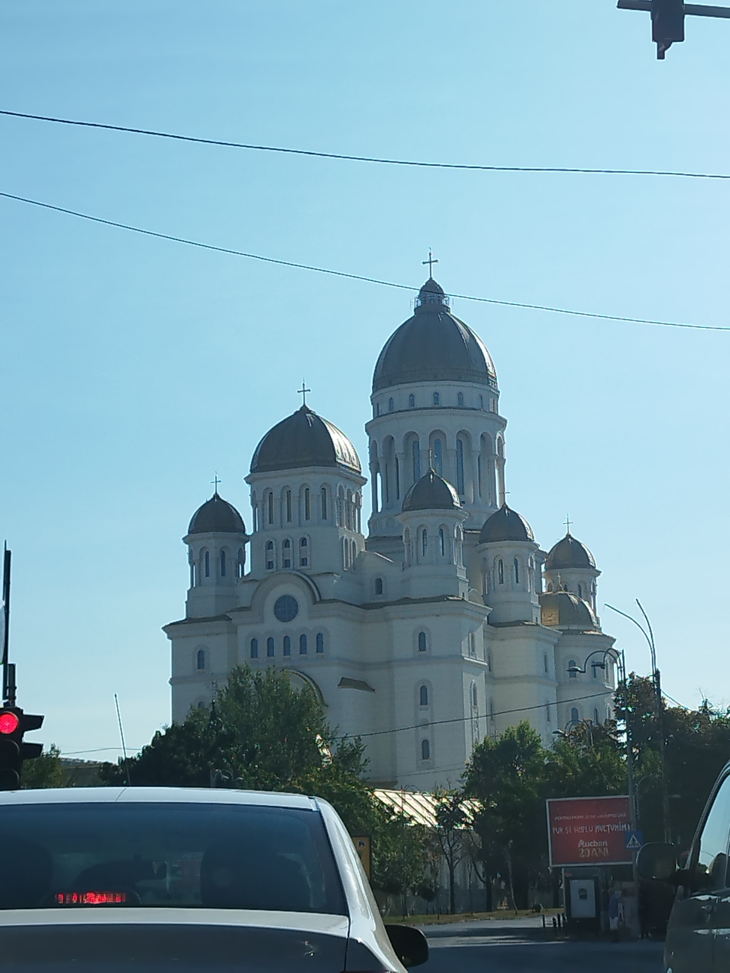 A large, white Orthodox cathedral with multiple golden domes and crosses is the central subject of the image, set against a clear blue sky. The cathedral has a classical architectural style with arched windows and ornate detailing. In the foreground, the rear of a white car is visible, suggesting the photo was taken from another vehicle. Traffic lights, with a red light illuminated, and street lamps are also present. Trees and a billboard with Romanian text and the Auchan logo are visible to the right of the cathedral. The scene appears to be a street in Bucharest, Romania, likely in front of the Cathedral of Saint Demetrius (Catedrala Sfântul Cuvios Dimitrie Basarabov).