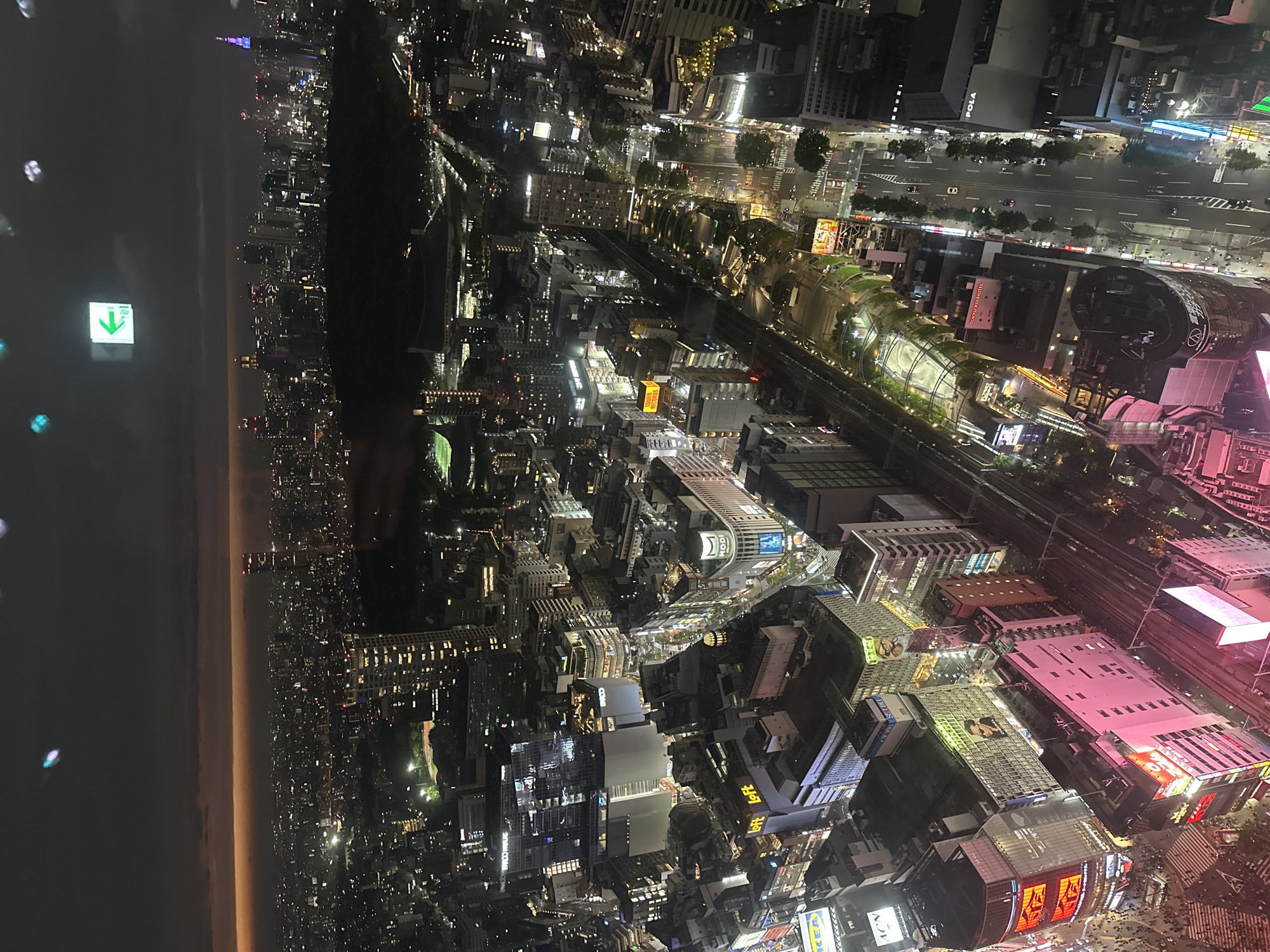 An aerial view captures the vibrant city of Shibuya, Japan, at night. The urban landscape is dominated by brightly lit buildings, illuminated streets, and distinct structures. The Tokyo Plaza can be identified by its unique cylindrical design and illuminated signage. Pedestrians and cars are visible on the busy streets, adding to the dynamic atmosphere. An exit sign is visible, as well as city lights extending to the horizon.