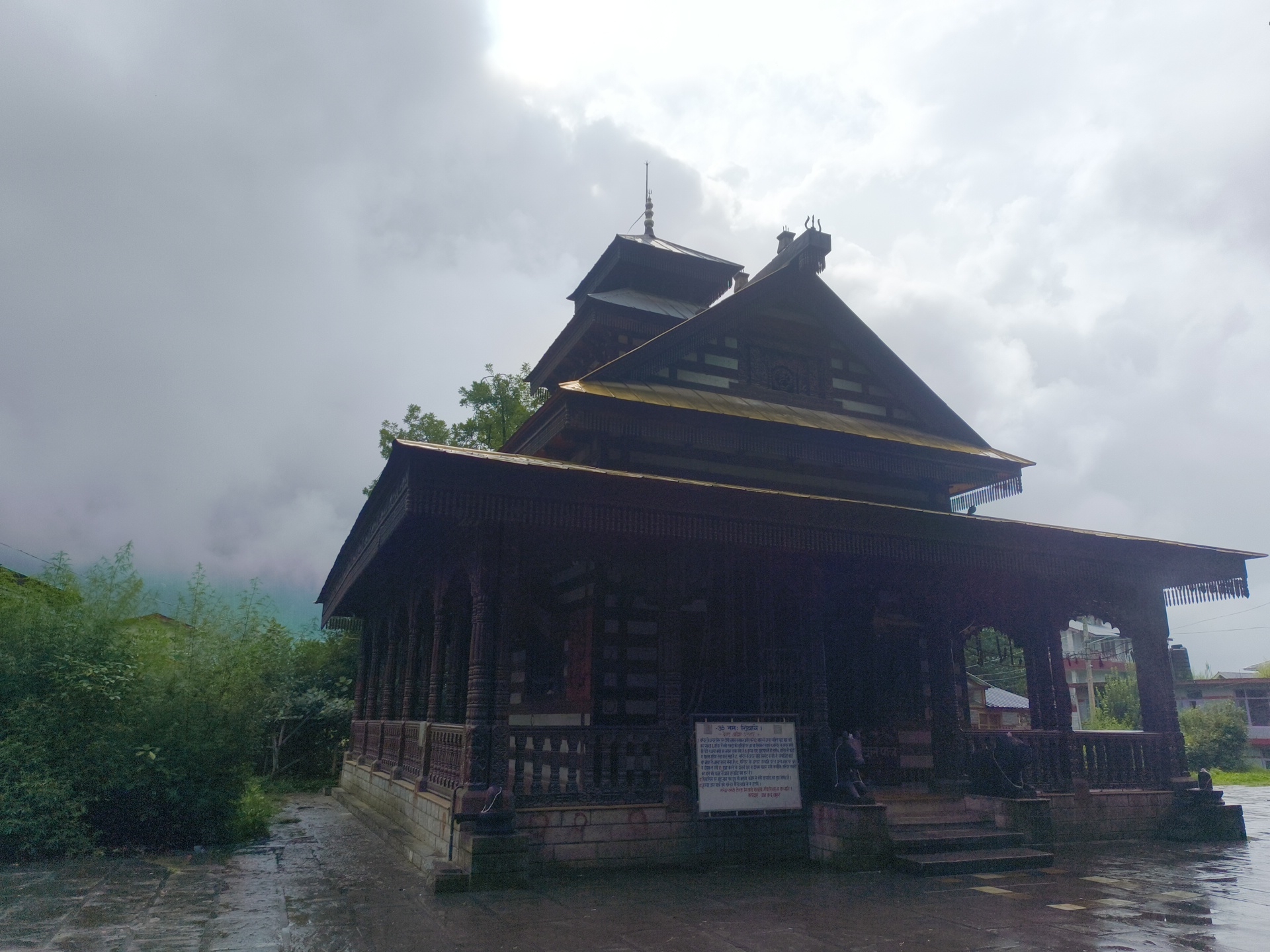 The image shows the Hadimba Devi Temple in Manali, India, on a cloudy day. The temple is a wooden structure with multiple tiered roofs and intricate carvings. A paved walkway, wet from rain, leads to the entrance of the temple, which features a wooden railing and decorative pillars. There is a sign with text written in Hindi, placed near the entrance. Lush green bushes and trees are visible to the left of the temple. In the background, there are some buildings and more greenery under a cloudy sky.